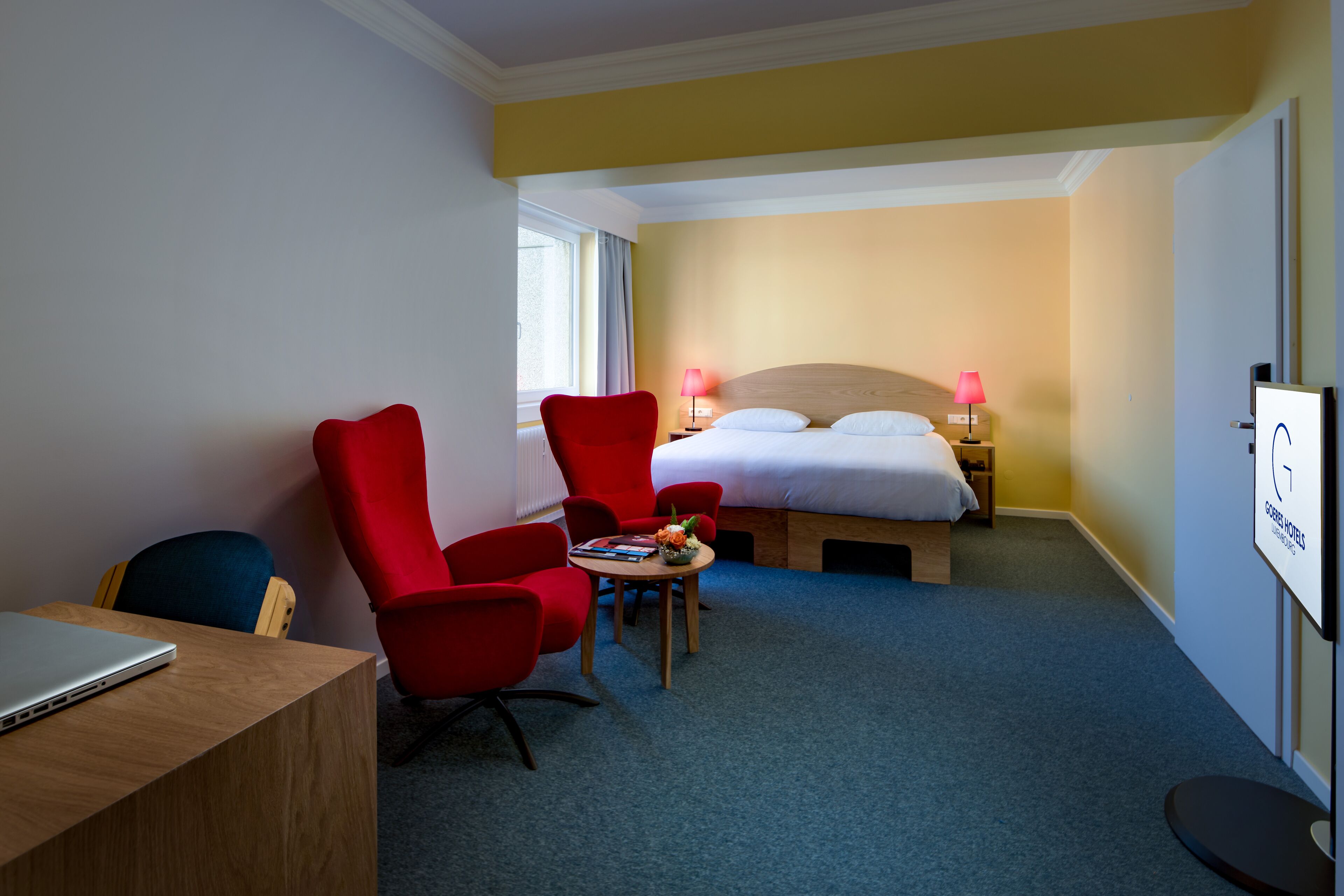 A hotel room with a bed, two red chairs, a small table with a vase, a laptop on a desk, and a TV on the wall.