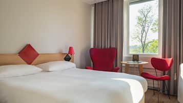 A hotel room with a large bed, two red chairs, a small table, and a window with curtains.
