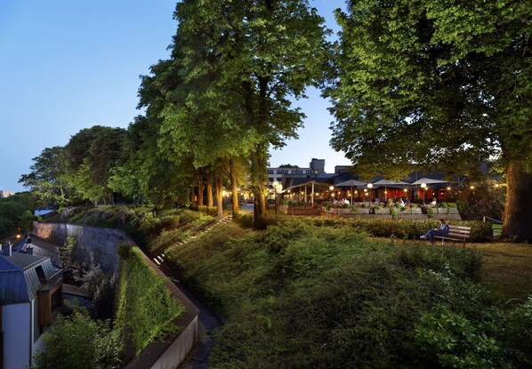 A lush, green outdoor area with a seating area and a building with multiple levels and outdoor lighting. - Hotel Parc Belle-Vue (Luxembourg City)