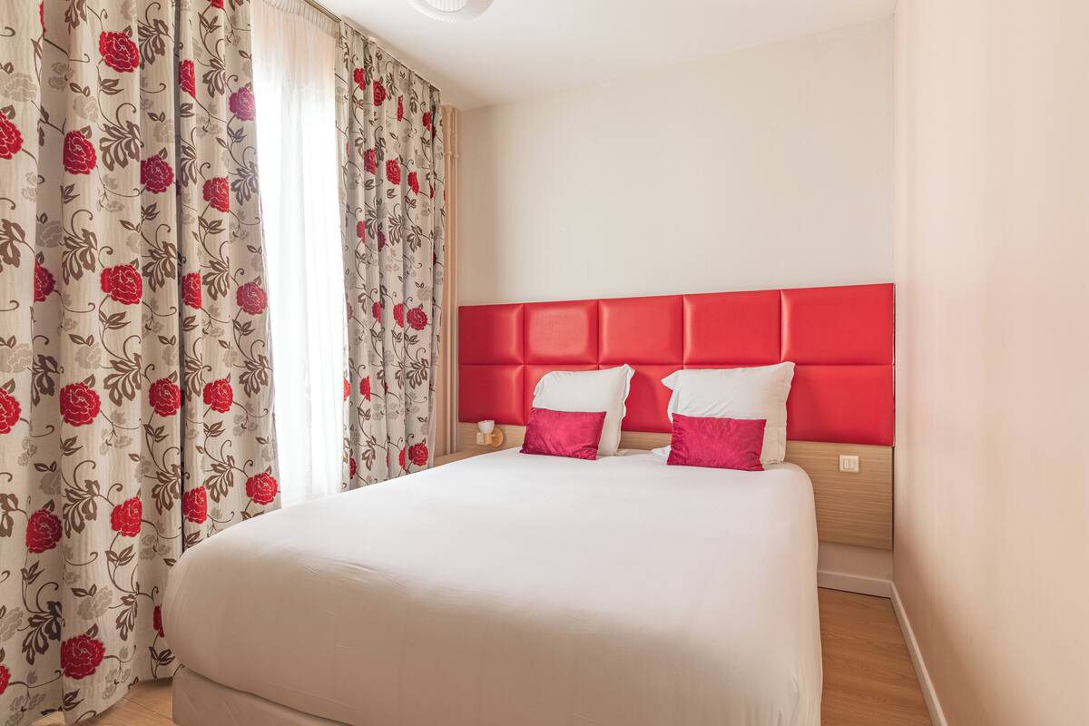 Habitación de hotel con una cama grande, cabecera roja, cortinas estampadas y mesita de noche de madera.