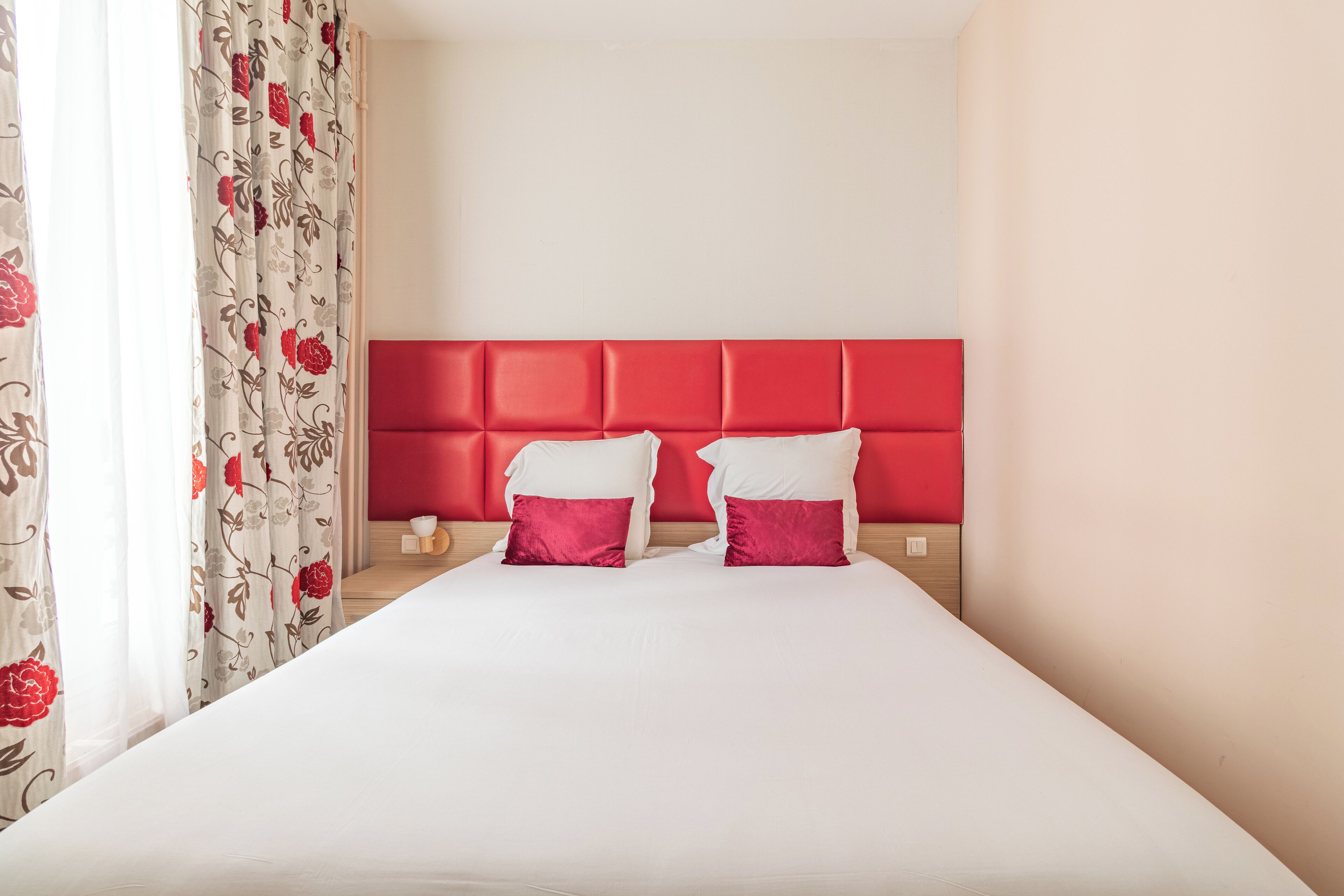 A hotel room with a large bed, red headboard, white bedding, and floral curtains.