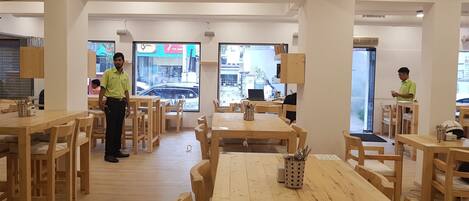 A restaurant interior with wooden tables and chairs, a person standing near the entrance, and a view of the street outside.