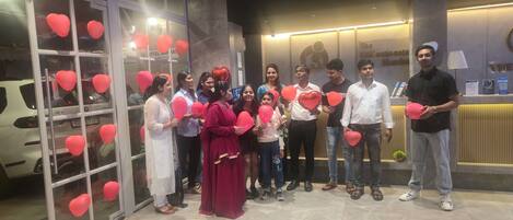 A group of people standing in front of a hotel reception area with a glass door, holding red heart-shaped balloons.