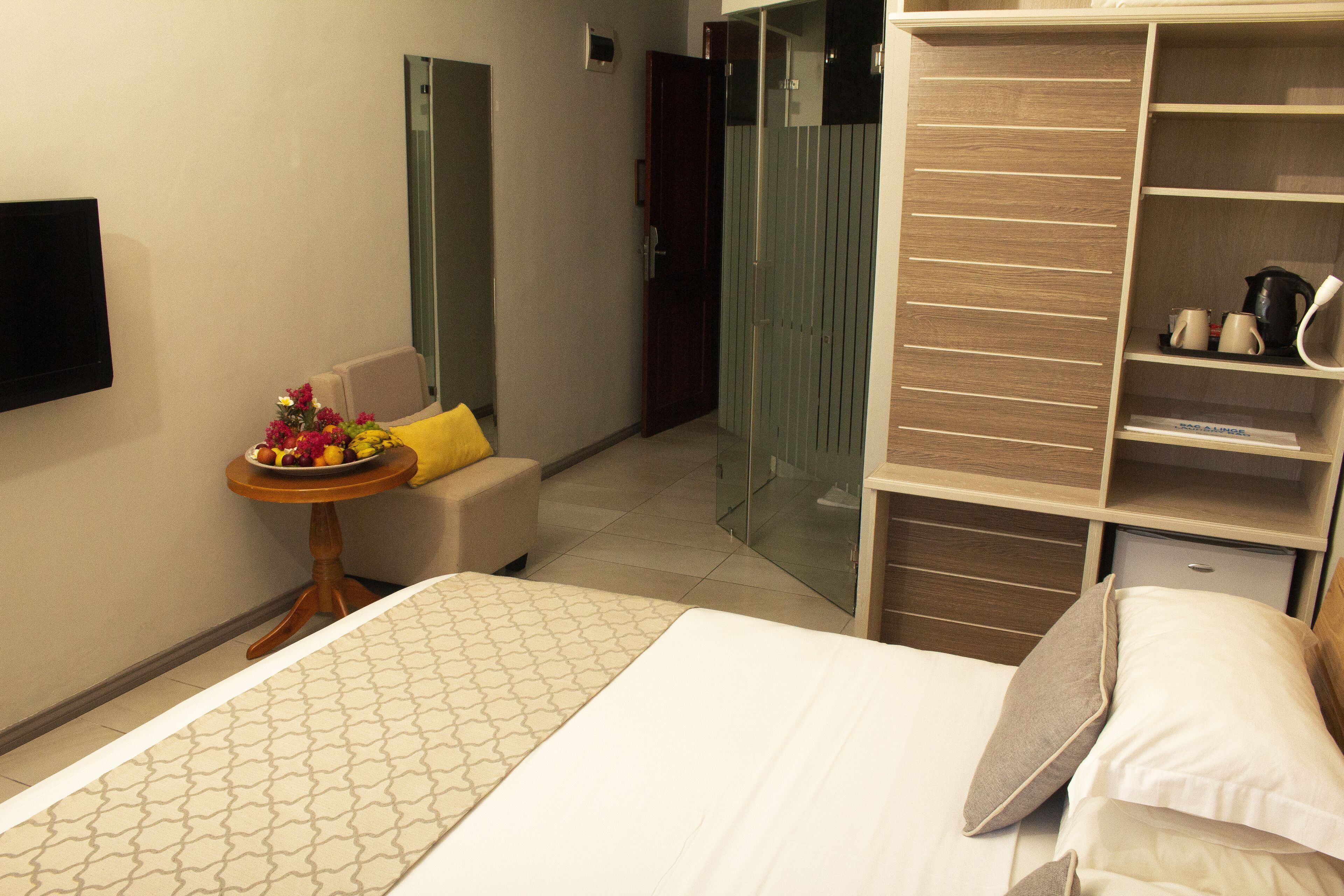 A hotel room with a bed, a small table with a fruit bowl, a chair, a wardrobe, and a TV mounted on the wall.