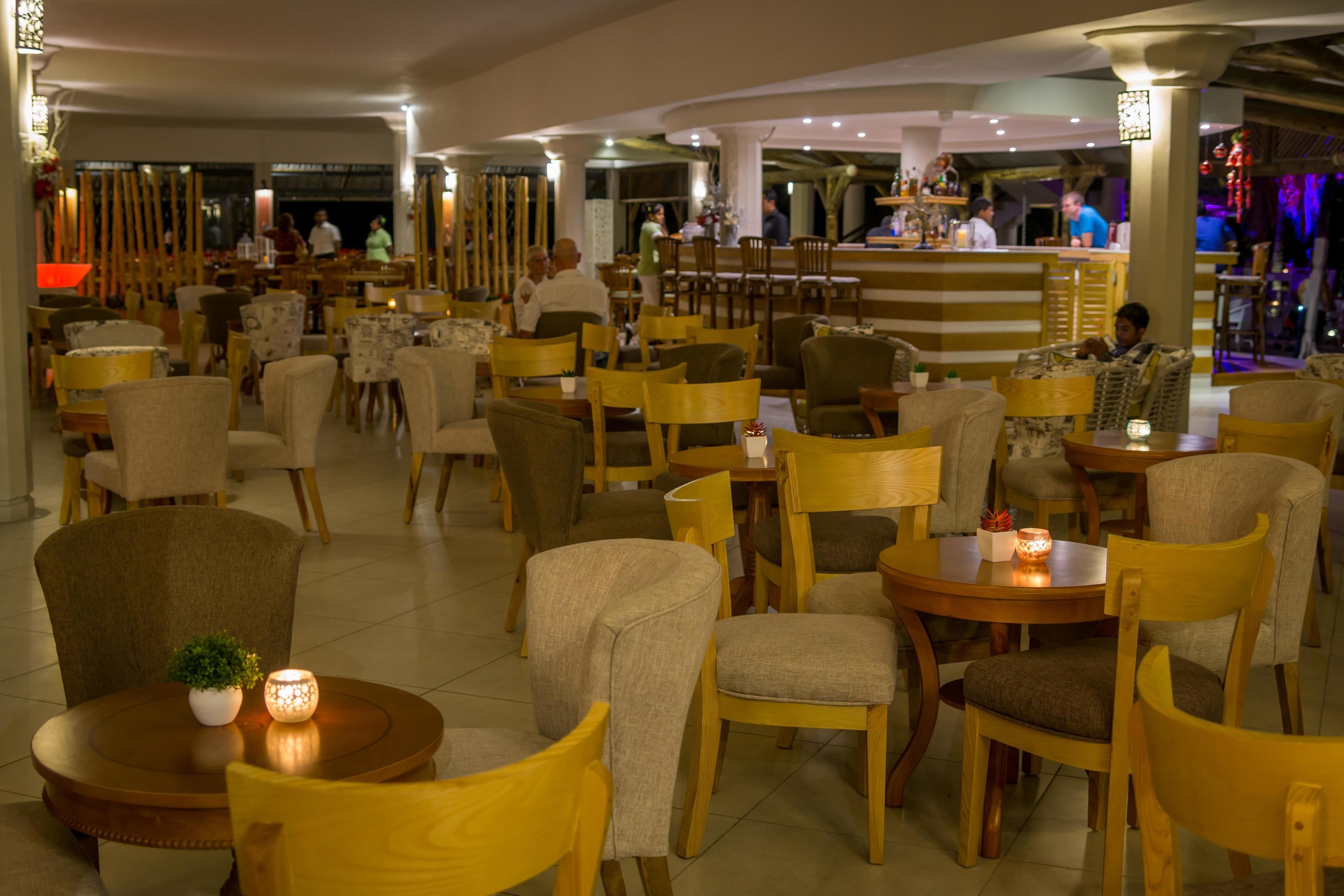 Un amplio comedor interior con varias mesas y sillas de madera, una zona de bar al fondo, y algunas personas cenando.
