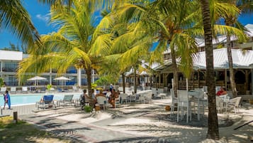 Un espace piscine aménagé dans un complexe hôtelier, avec des chaises longues et des parasols, entouré de palmiers et bordé d’un bâtiment au toit blanc.