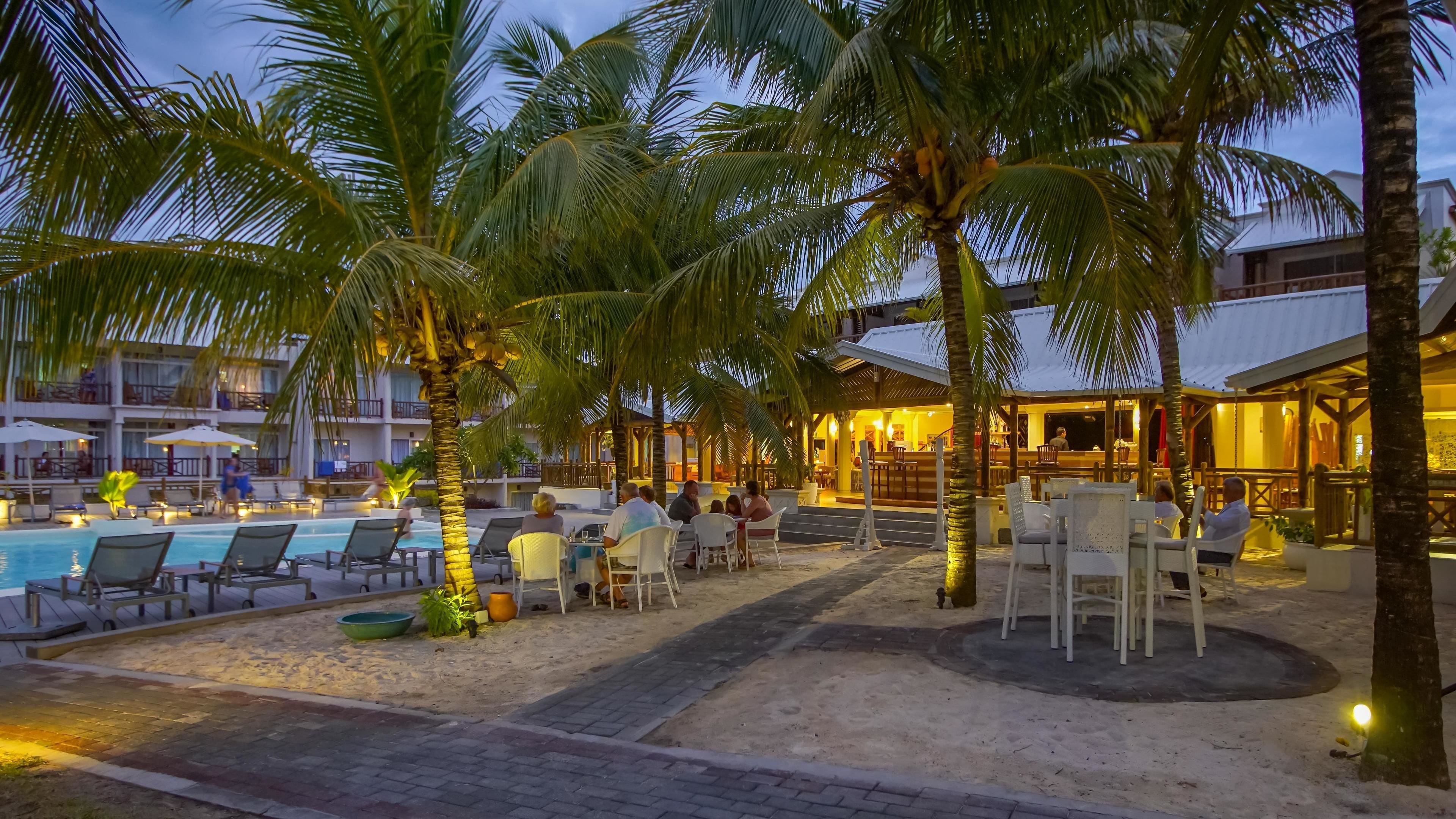 Un área de piscina en un resort, con sillones de descanso y sombrillas, rodeada de palmeras y un bar o área de lounge.