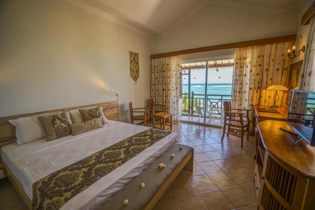A hotel room with a large bed, a wooden bench, a TV, and a balcony with a view of the sea.