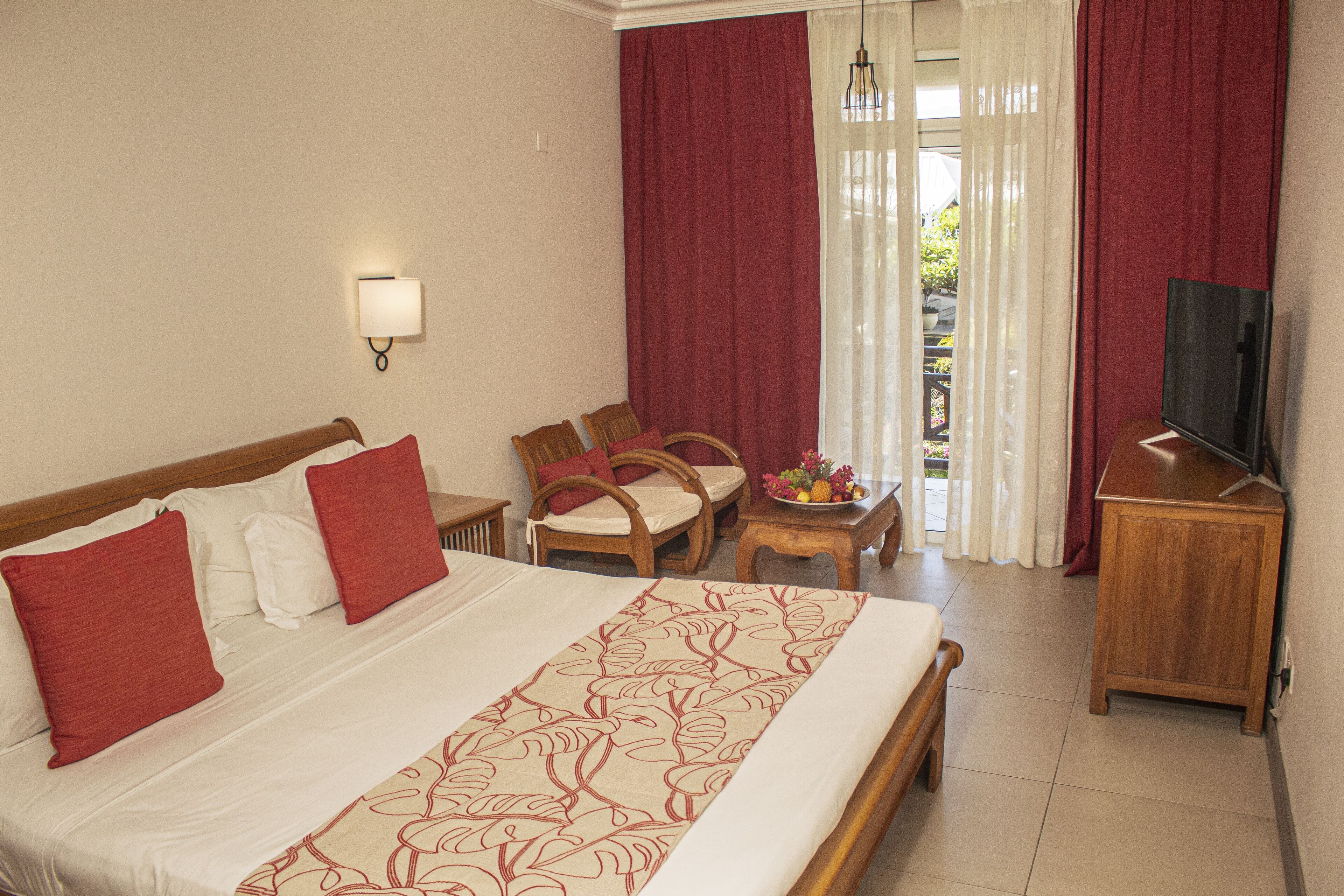A hotel room with a large bed, two chairs, a small table with a fruit bowl, a TV, and a view of the outdoors through sheer curtains.