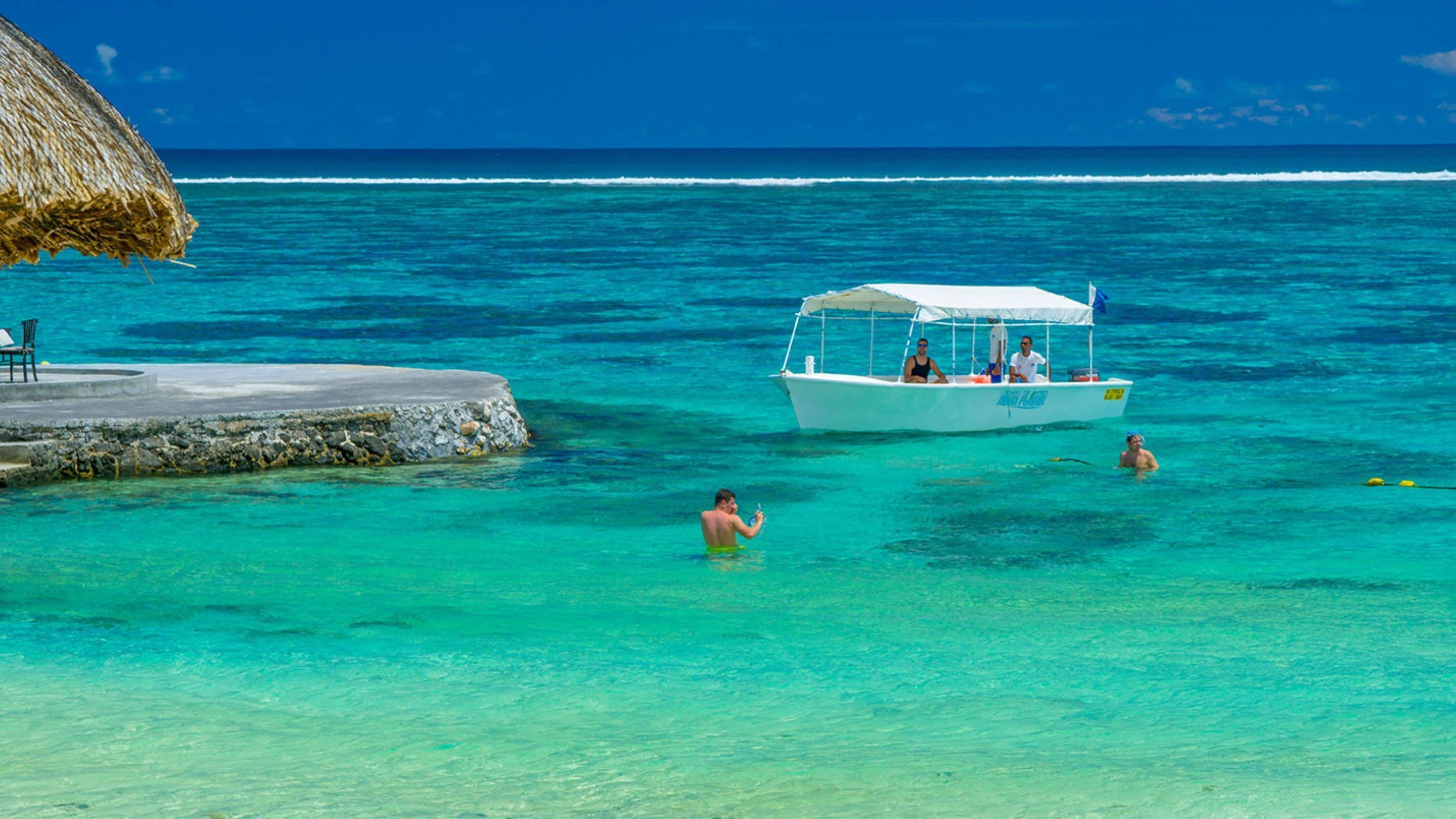 A boat with passengers in clear turquoise waters, a dock with a thatched roof, and a person swimming.
