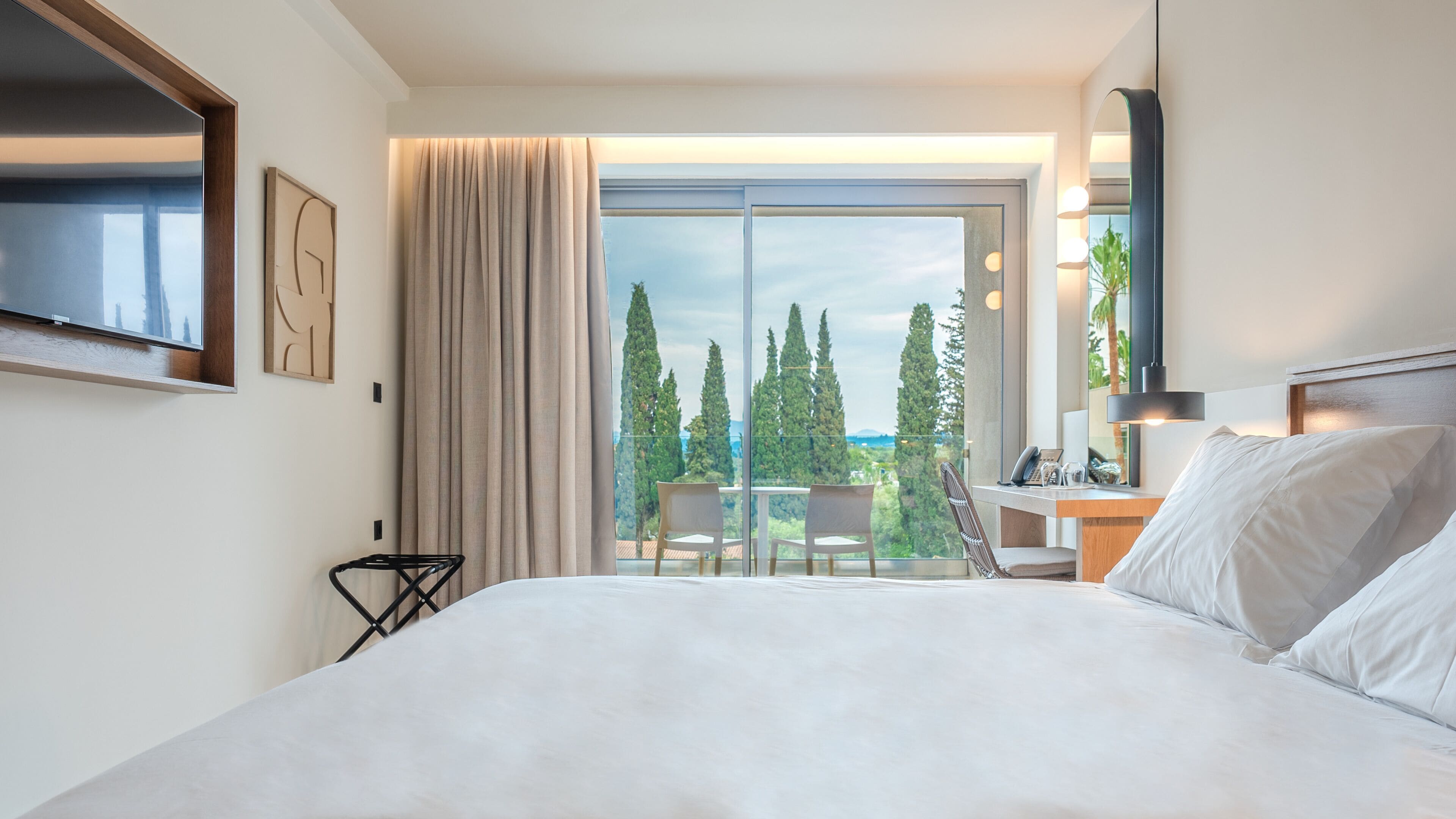 Een hotelkamer met een groot bed, een bureau met stoel, een aan de muur bevestigde televisie en uitzicht op bomen en een balkon.