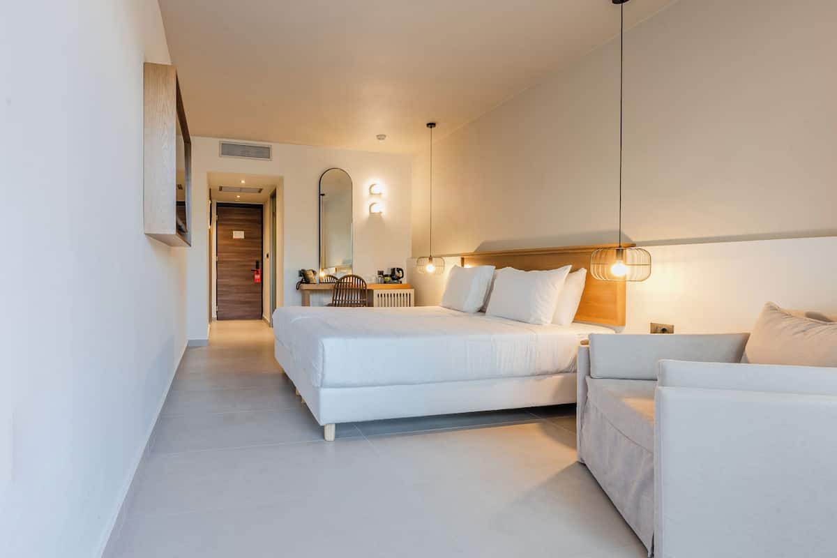 A modern hotel room with a large bed, a sofa, a desk, and a mirror.