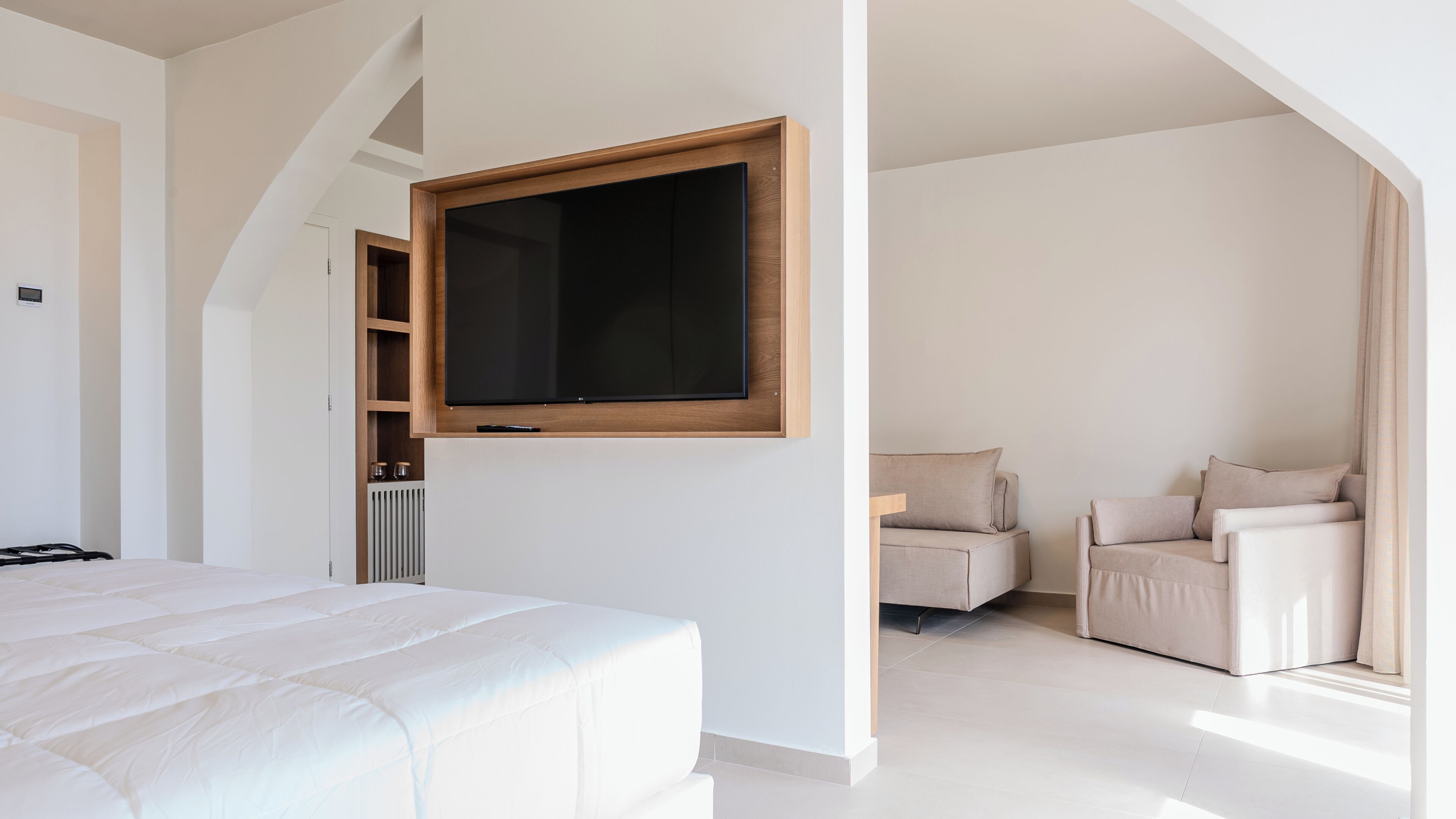 A modern bedroom with a large bed, a TV mounted on the wall, a seating area with a sofa and armchairs, and a radiator.