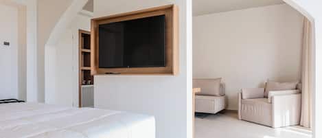 A modern bedroom with a large bed, a TV mounted on the wall, a seating area with a sofa and armchairs, and a radiator.