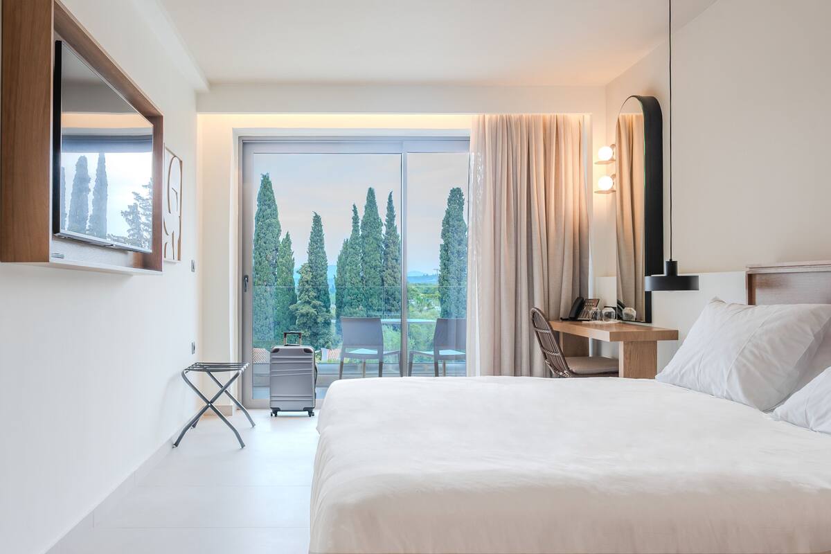 A modern hotel room with a large bed, a desk with a lamp, a chair, a small table with a phone and glass, and a view of trees through the window.