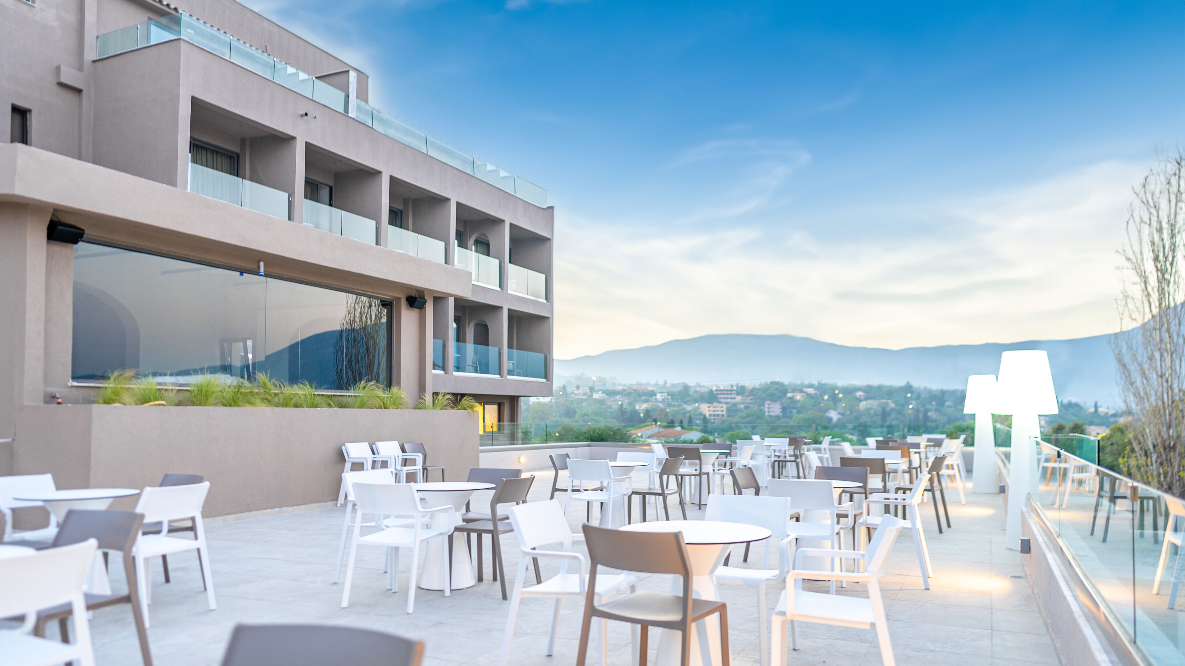 A rooftop terrace with outdoor seating, a modern building with large windows, and a view of distant hills.