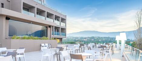 A rooftop terrace with outdoor seating, a modern building with large windows, and a view of distant hills.