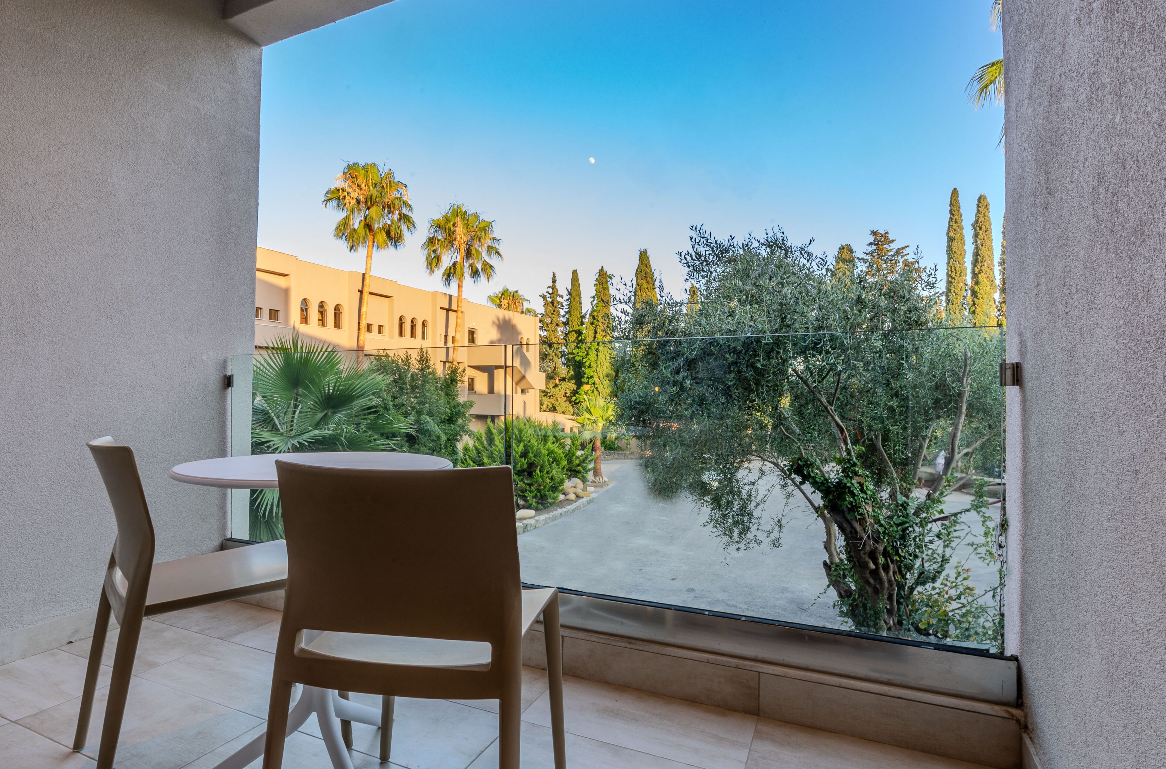 Un balcón con mesa y sillas, con vista a un patio con palmeras y un edificio al fondo.