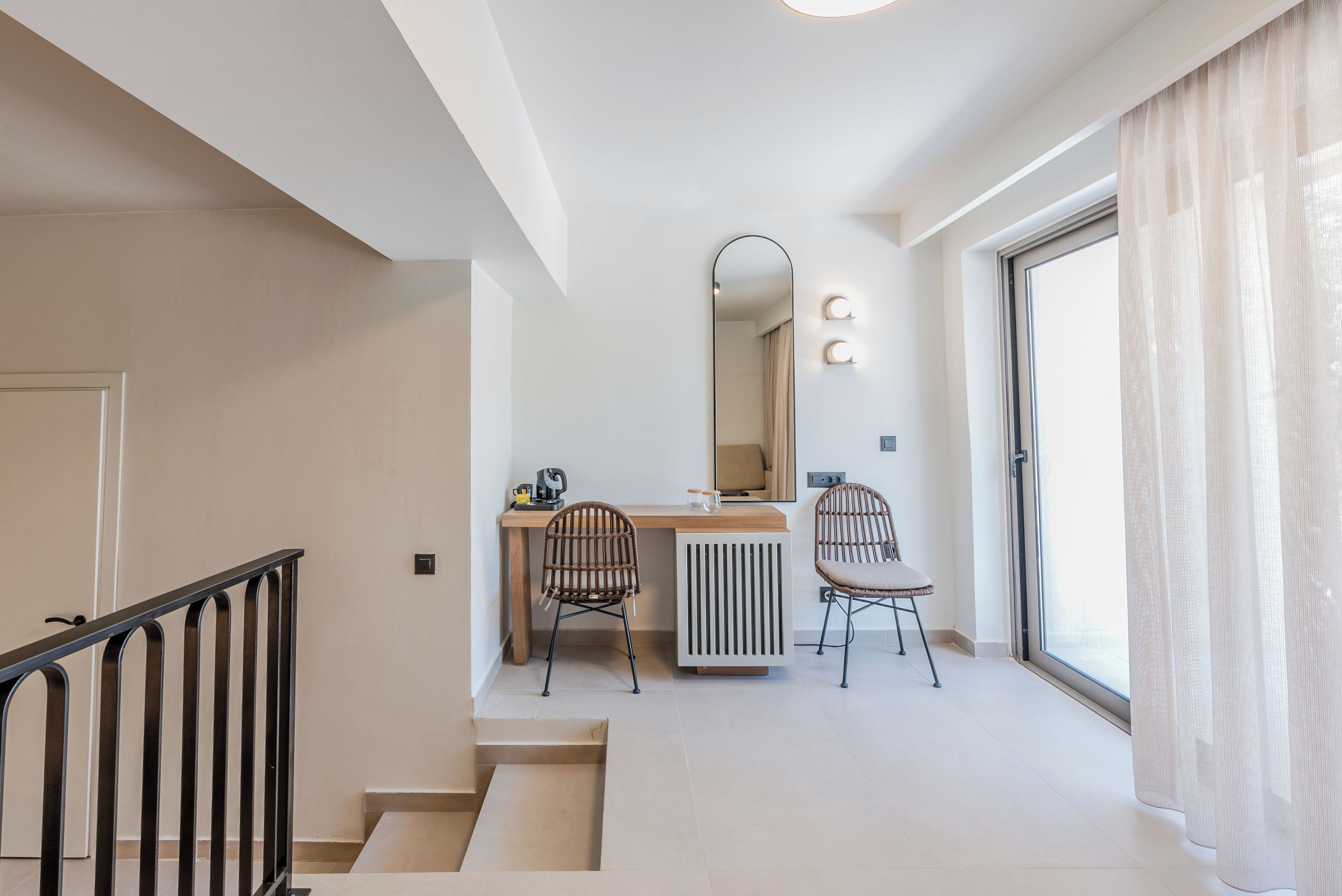 Un interior modern amb una escala, un escriptori amb dues cadires, un radiador i una finestra gran amb cortines.