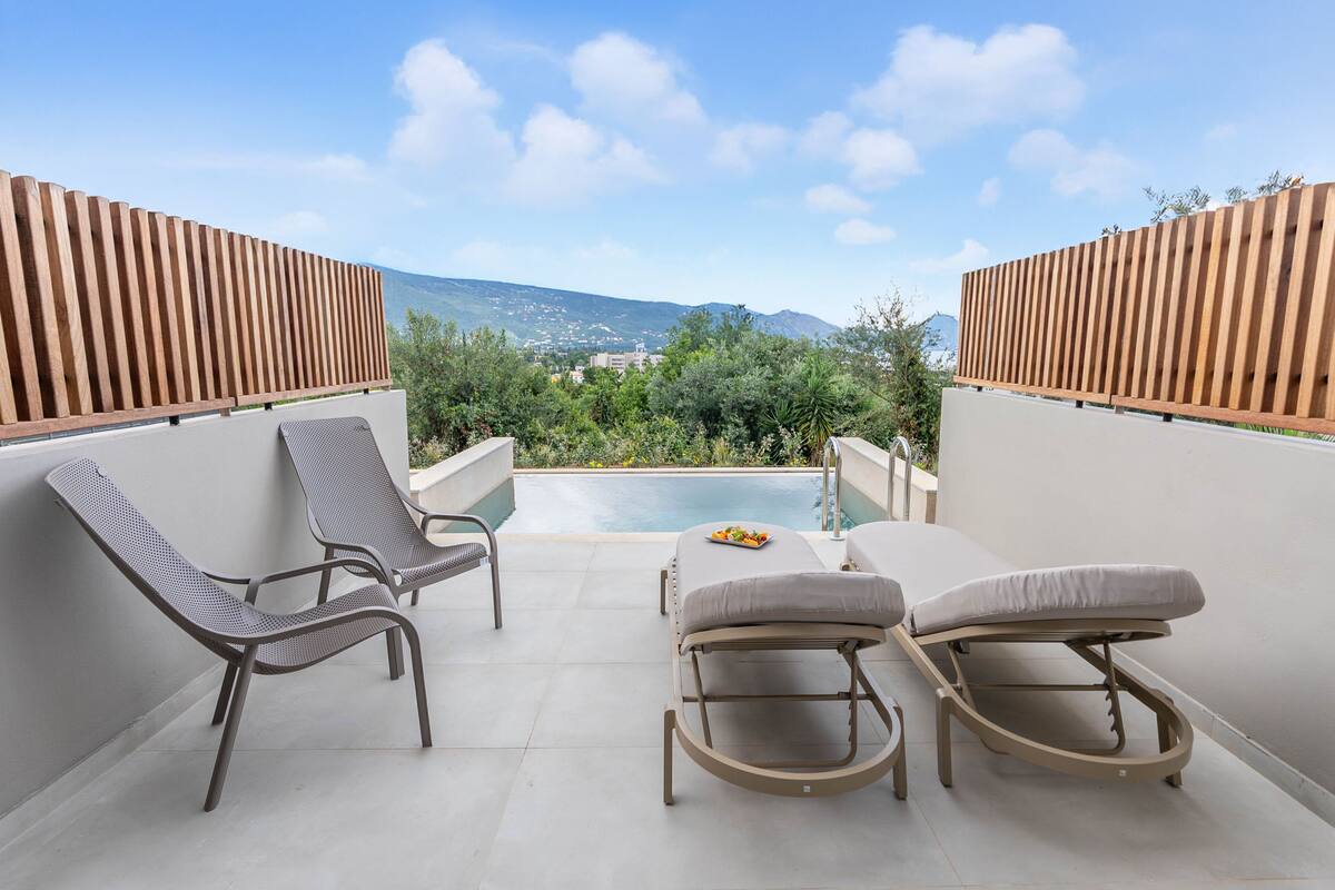 A balcony with two lounge chairs and a table, overlooking a swimming pool and a mountainous landscape.