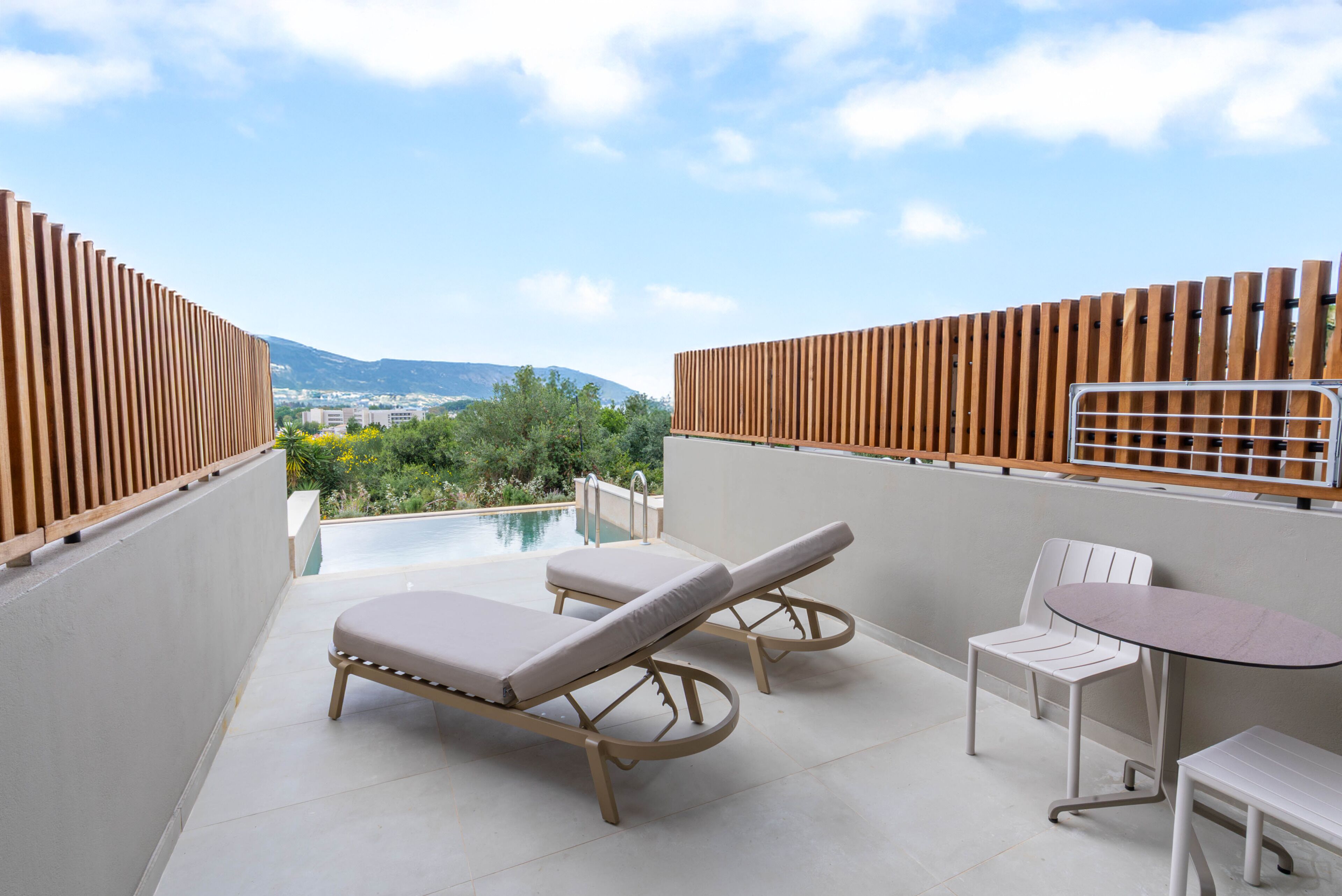 A rooftop terrace with two lounge chairs, a table, and a view of a swimming pool and distant landscape.