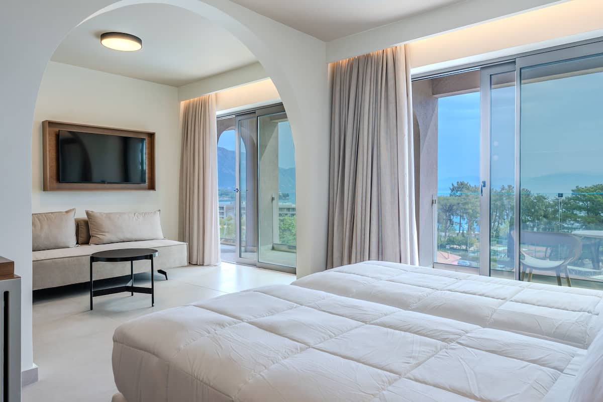 A modern bedroom with a large bed, a sofa, a TV mounted on the wall, and a balcony with curtains.