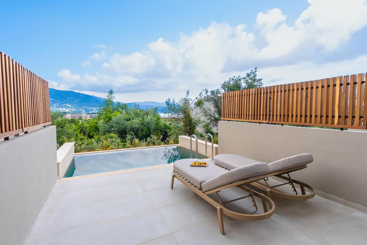 A sun lounger with a cushion and a tray of fruit on a tiled patio with a pool and a view of greenery and mountains.