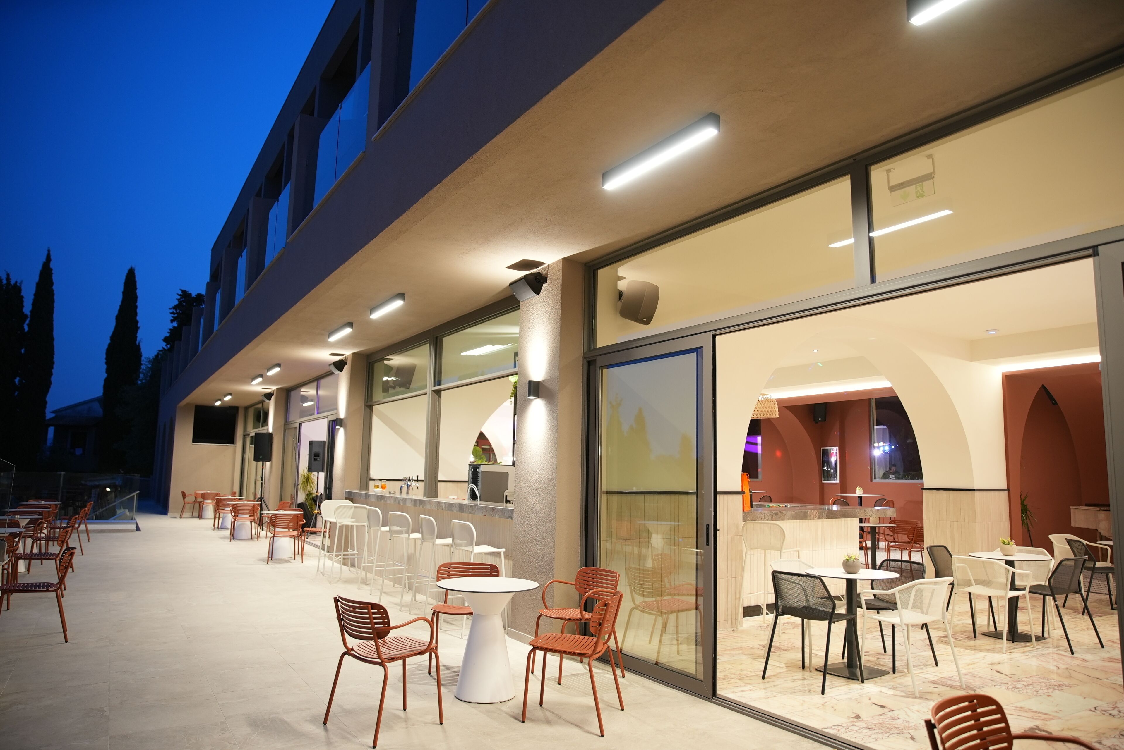Un'area pranzo all'aperto con tavoli, sedie e un bar, separata dall'interno da un muro di vetro.