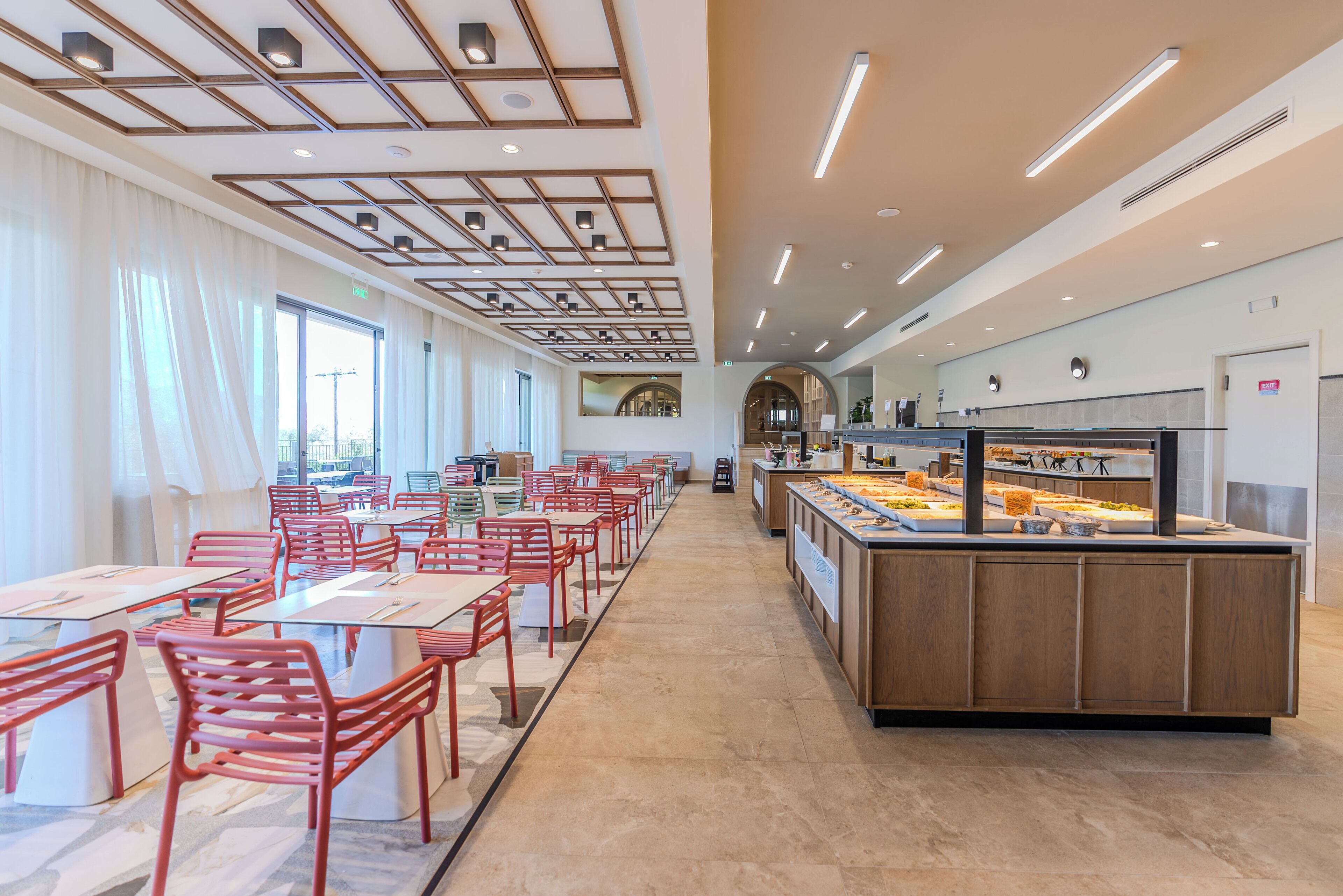 A spacious restaurant with long tables and chairs, a central counter with a display case, and a modern ceiling design.
