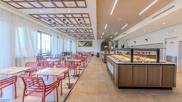 A spacious restaurant with long tables and chairs, a central counter with a display case, and a modern ceiling design.