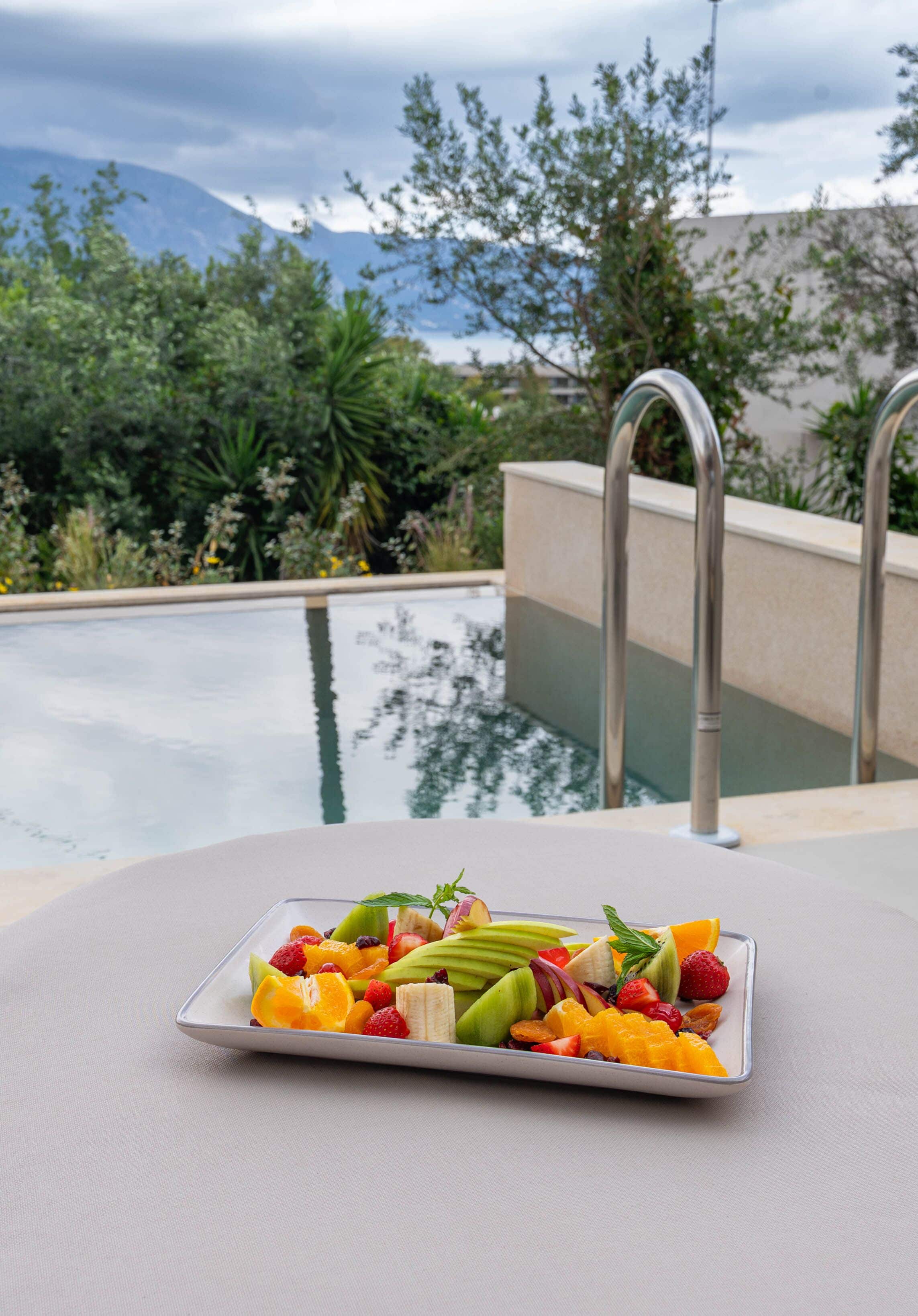 Una bandeja de frutas sobre una mesa junto a una piscina, con vistas a montañas y vegetación.