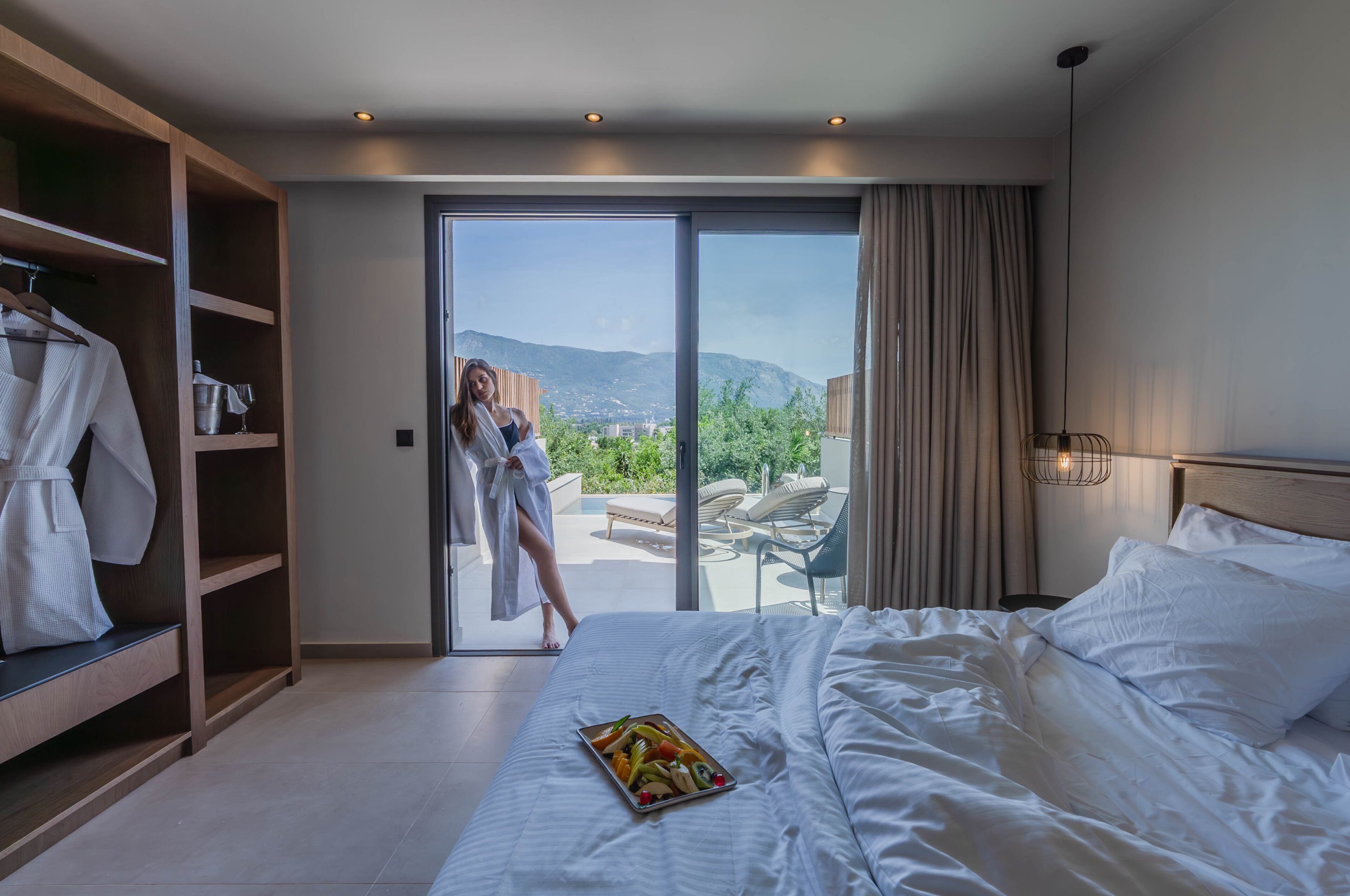 A hotel room with a bed, a tray of fruit, a wardrobe, and a view of the outdoors.