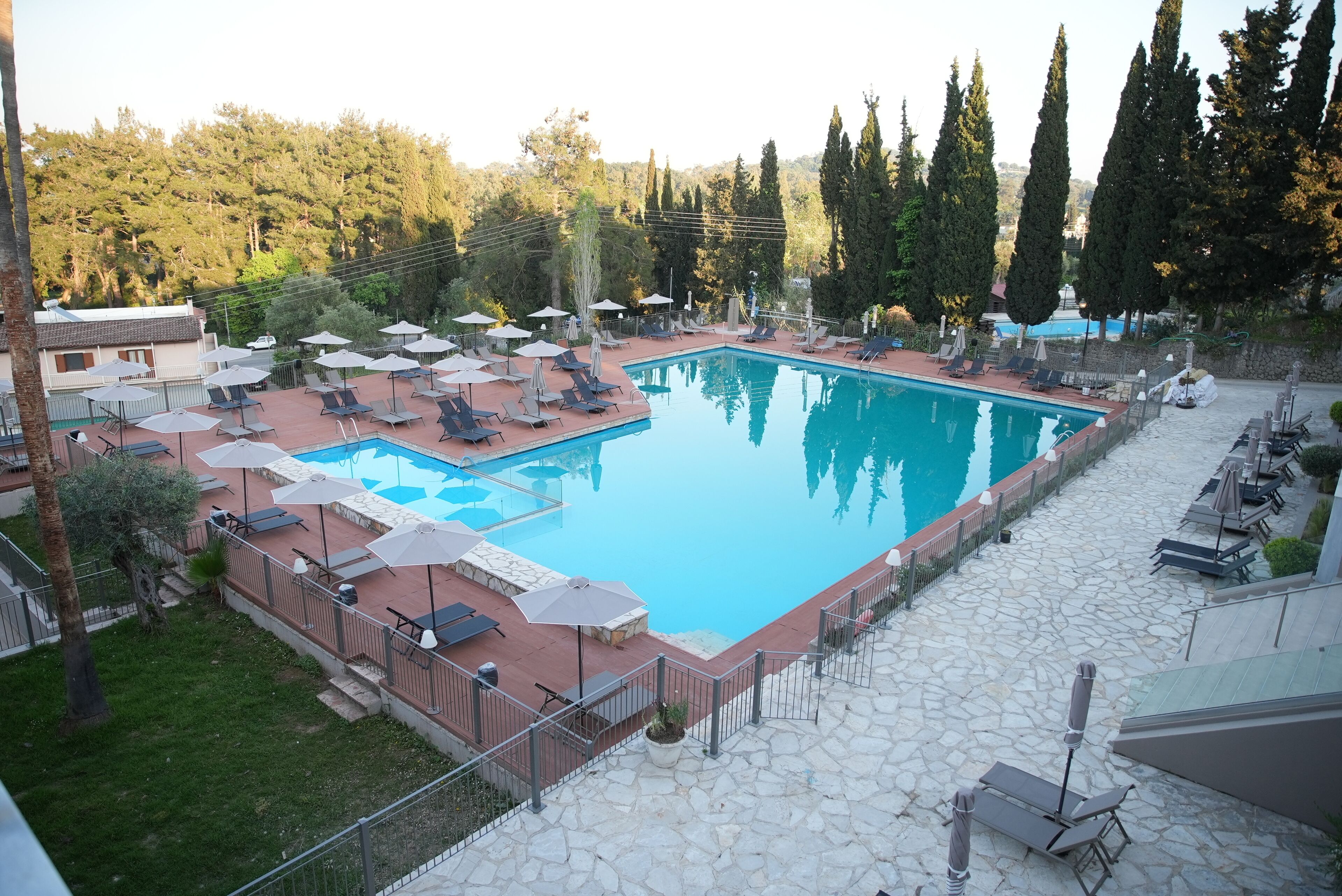 Una piscina rettangolare con lettini e ombrelloni, circondata da pavimento in pietra e una recinzione.