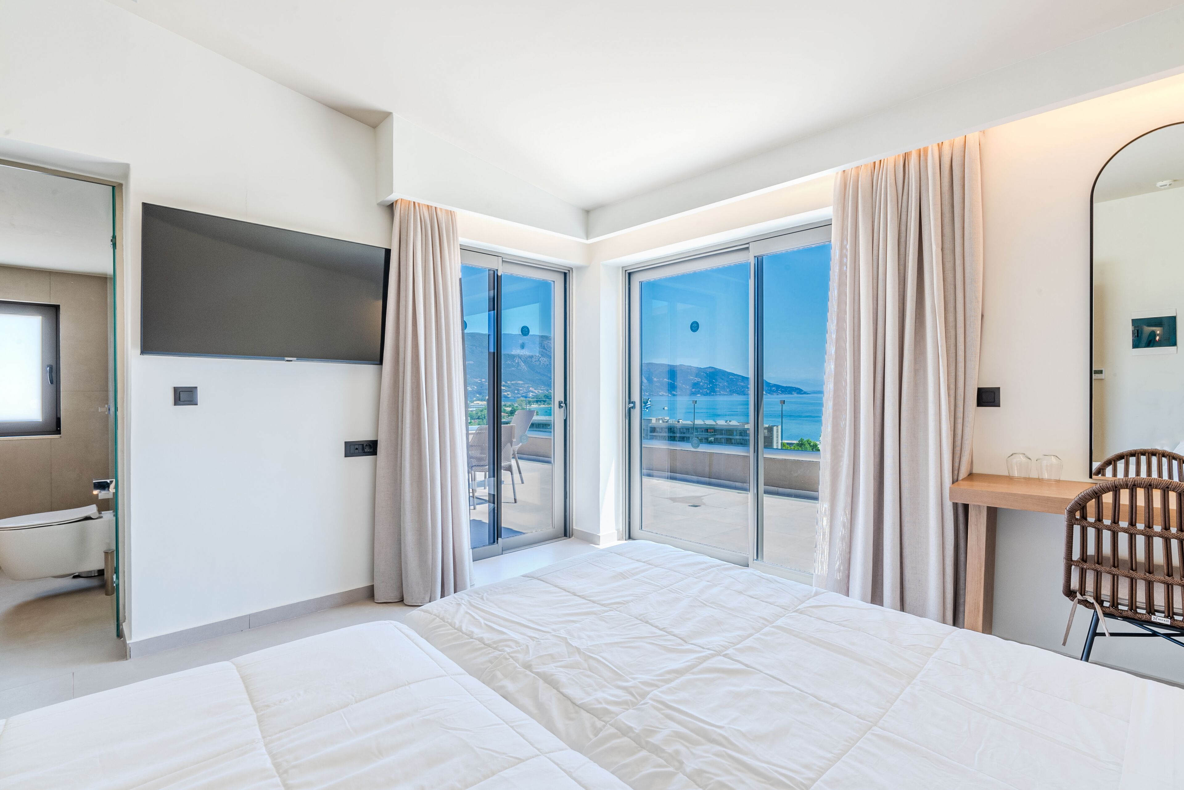 A hotel room with a large bed, a TV mounted on the wall, a bathroom with a toilet, and a balcony with a view of the sea.