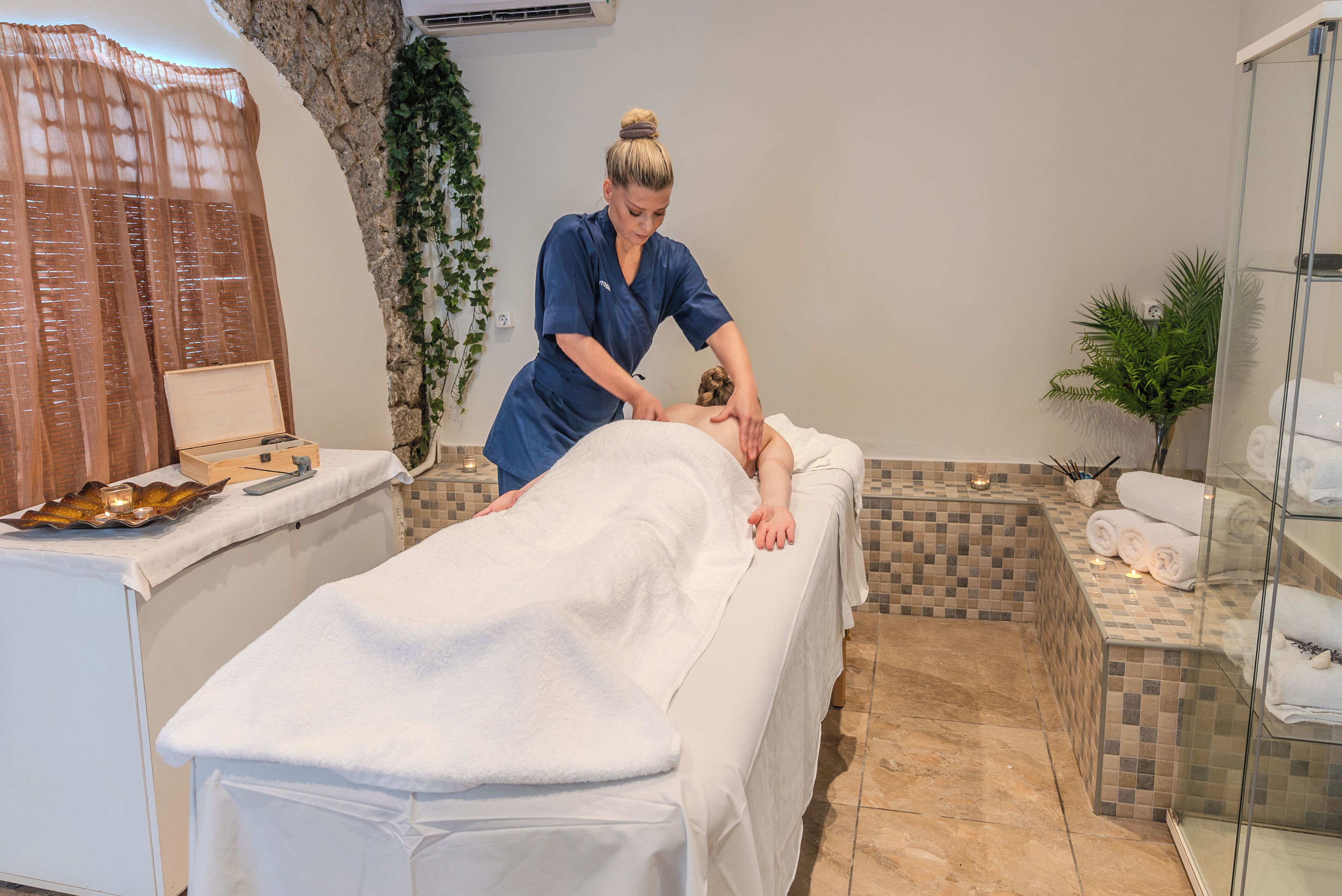 A massage therapy room with a massage table, a towel-covered client, and a therapist in blue scrubs.