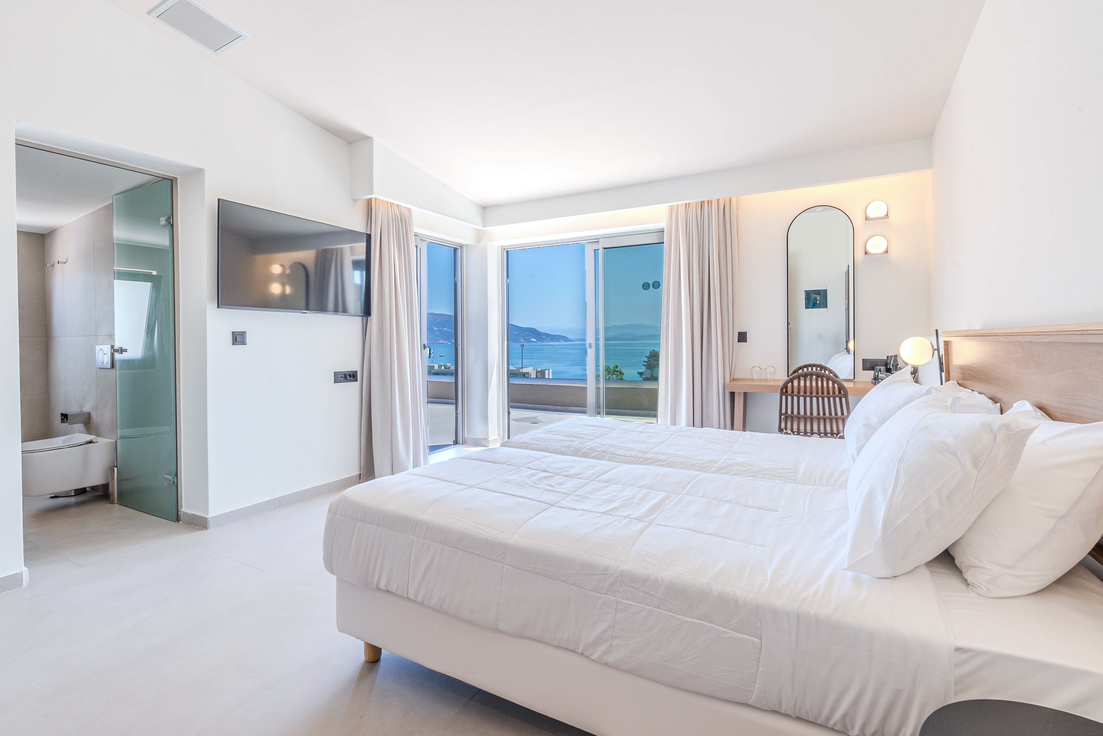 Una habitación de hotel moderna con una cama grande, un baño con puerta de vidrio y un balcón con vistas al mar.