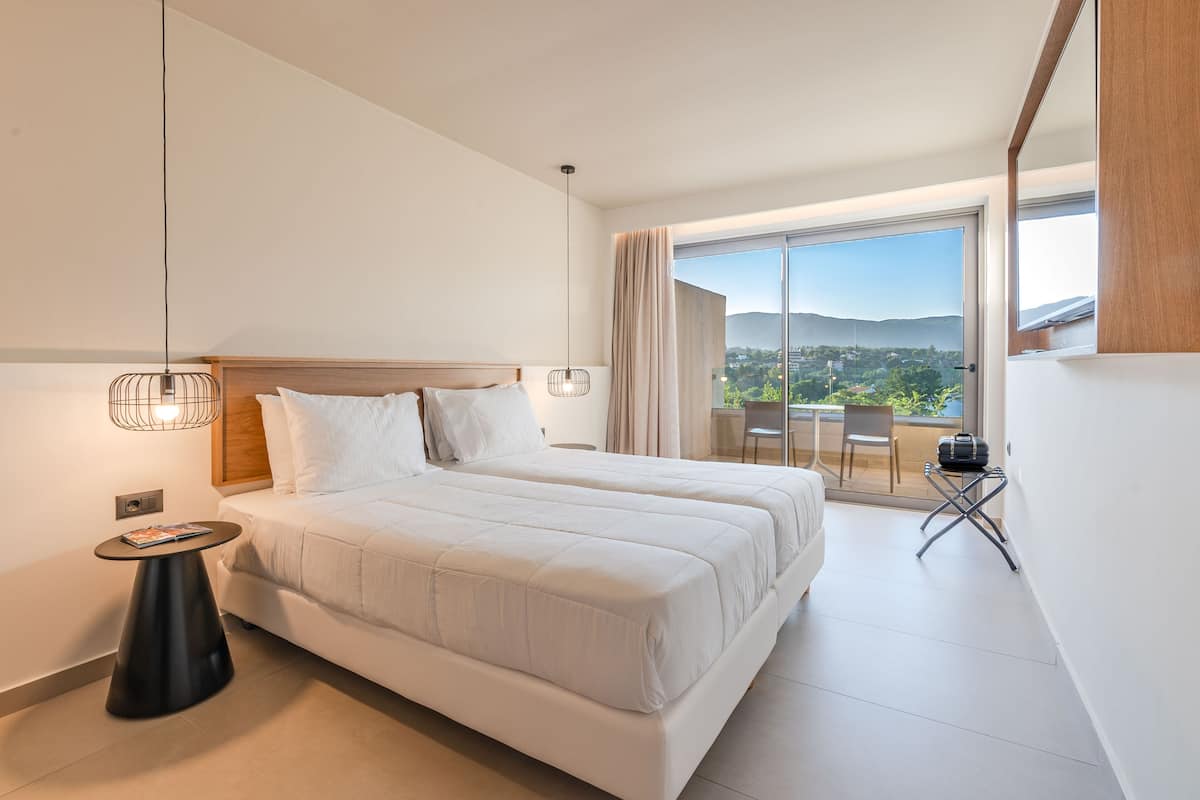 A modern hotel room with a large bed, a small table, and a balcony with a view of the city.
