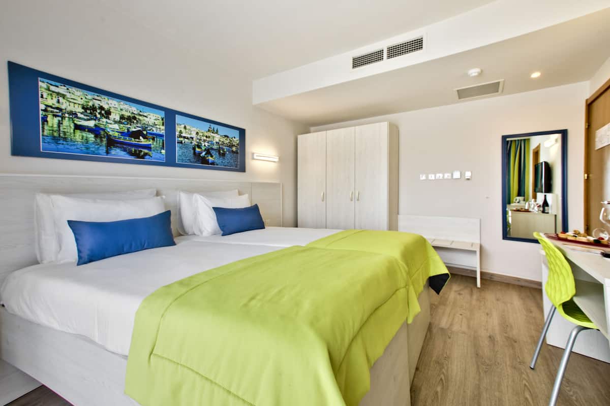 Quarto de hotel com cama, escrivaninha e cadeira. Há duas pinturas na parede e um guarda-roupa.