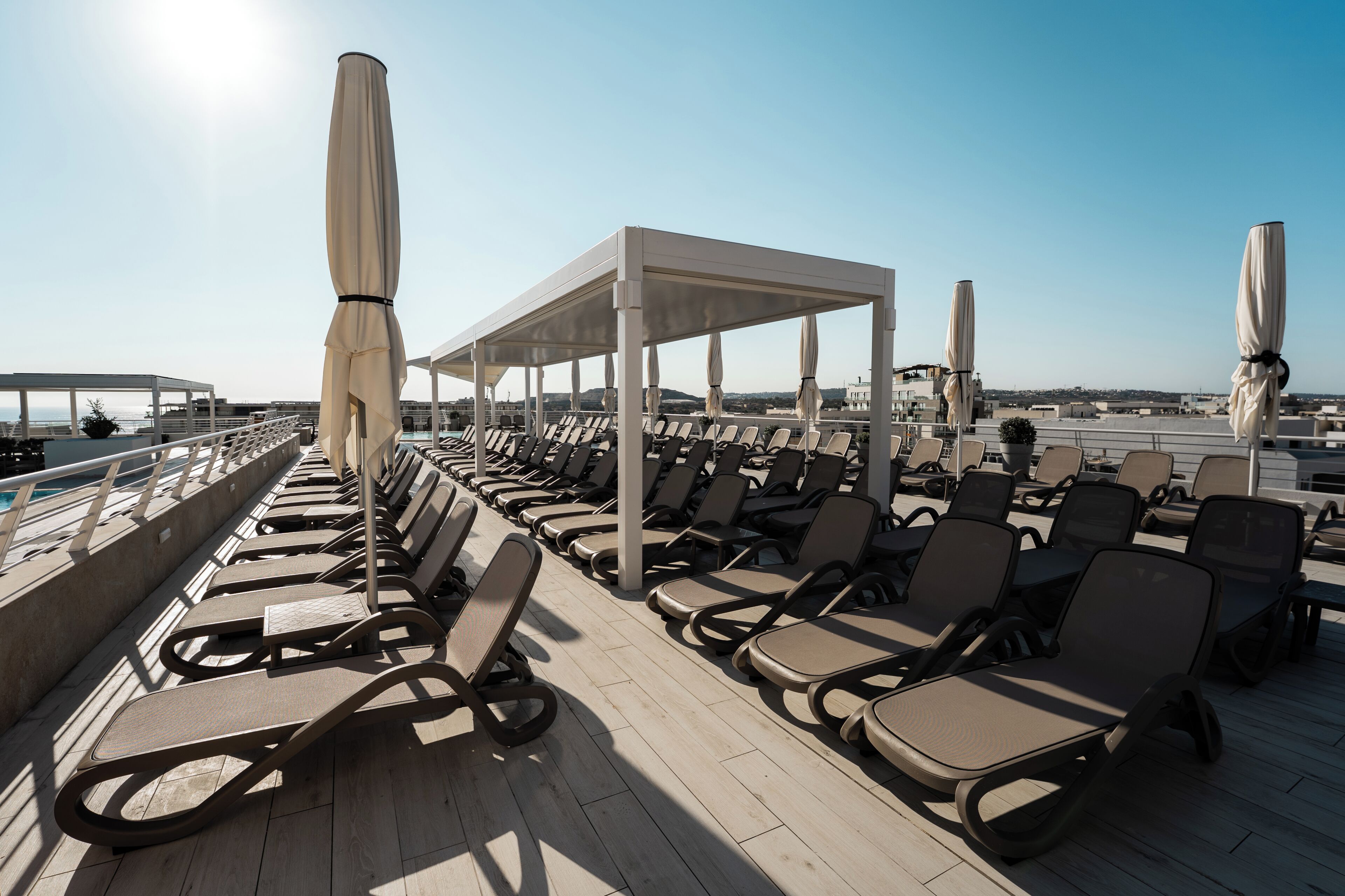 A sunlit outdoor seating area with rows of lounge chairs and umbrellas, overlooking a body of water.