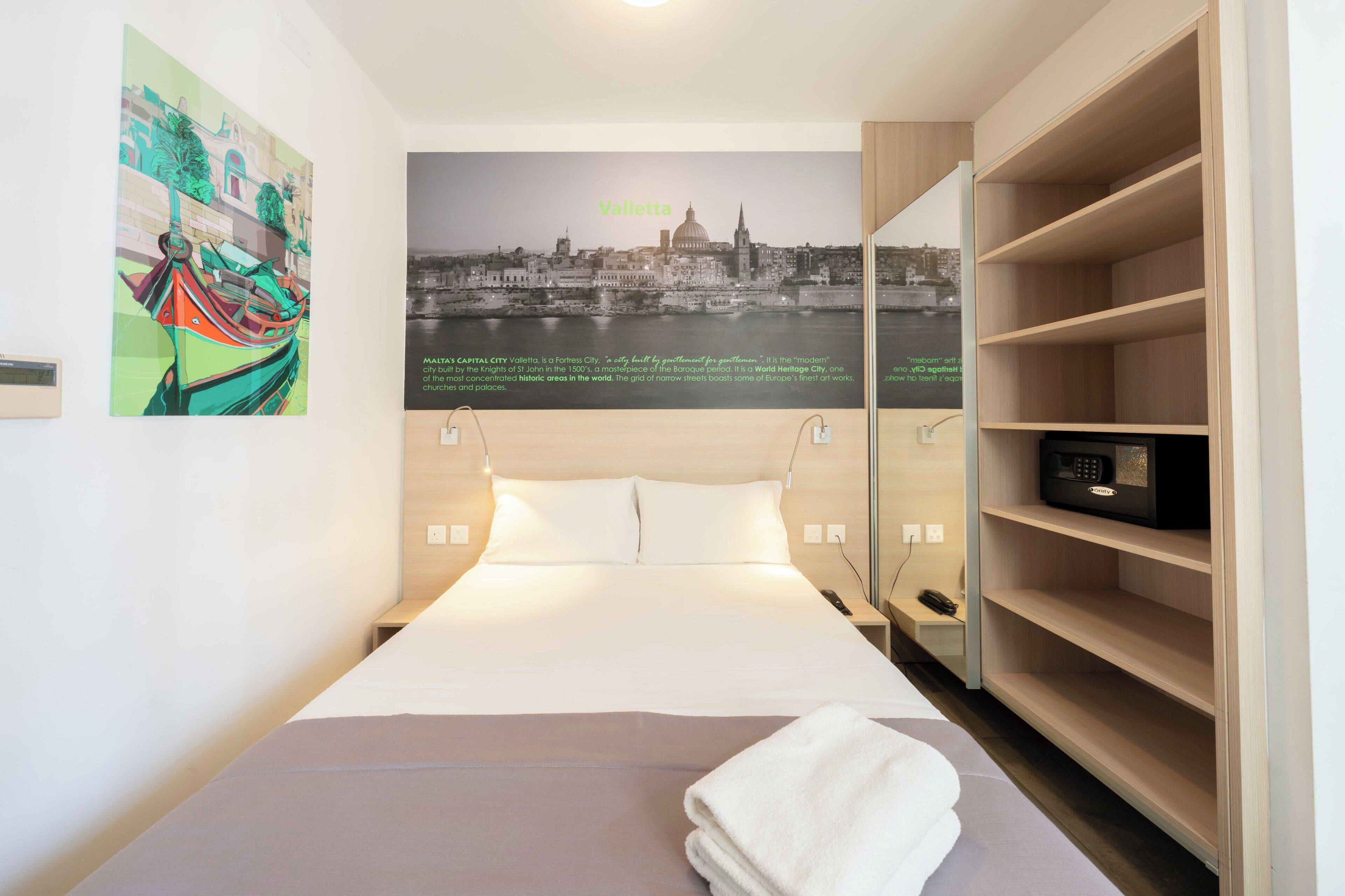 Une chambre d’hôtel avec un lit, un téléviseur fixé au mur, une étagère et une affiche représentant La Valette.
