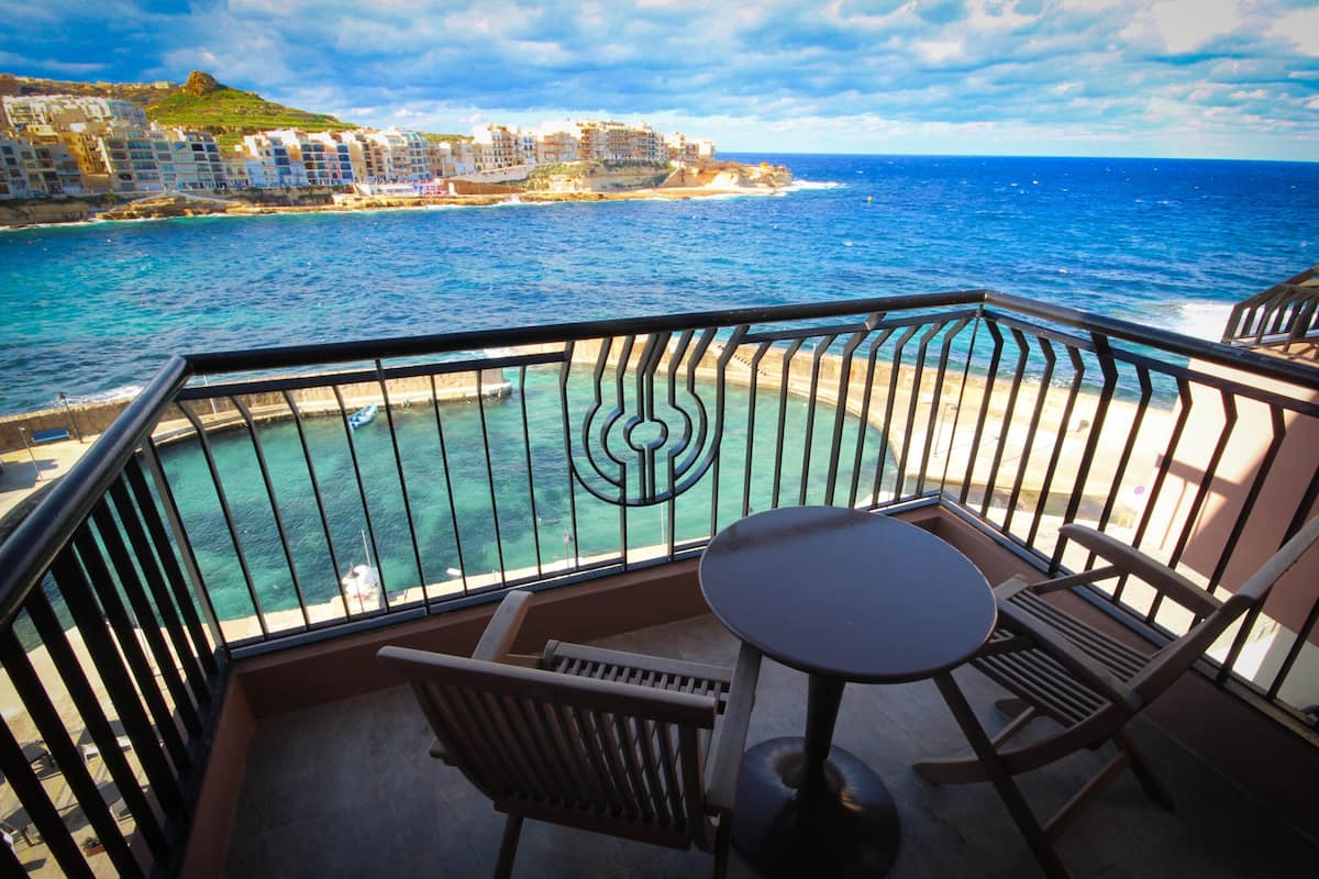 A balcony with a table and chairs, overlooking a coastal area with buildings and the sea.