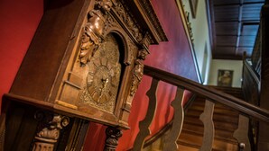 A detailed grandfather clock with ornate carvings, positioned near a wooden staircase with a metal handrail. - Hoftaferne Neuburg (Neuburg am Inn)