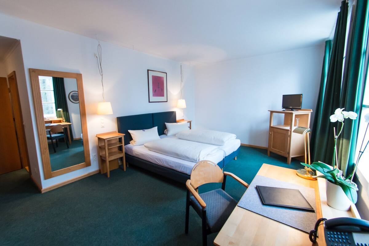 A hotel room with a bed, desk, chair, lamp, and a small table with a plant.