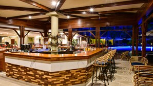 A bar area with a wooden counter, bar stools, and a pool area visible through the glass. - Tarisa Resort (Mont Choisy)