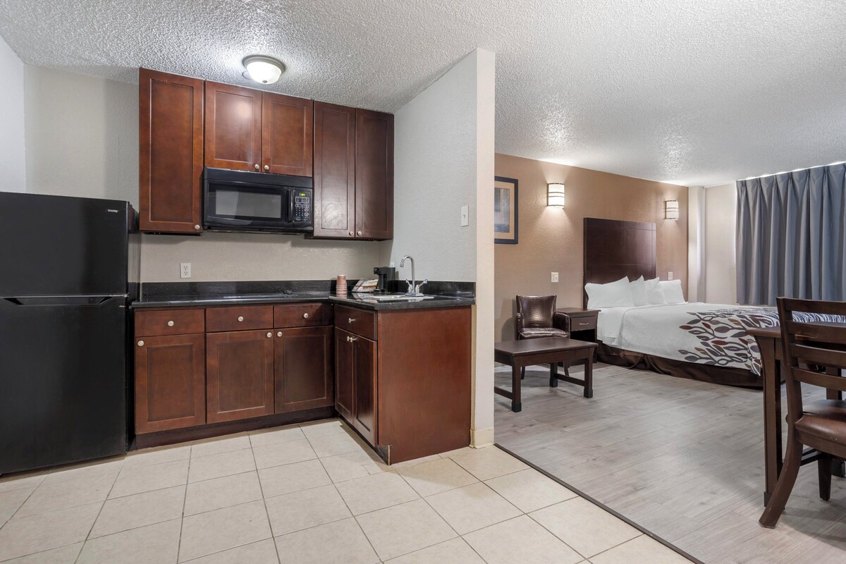 A compact hotel room with a kitchenette, a bed with a headboard, a desk, and a chair.
