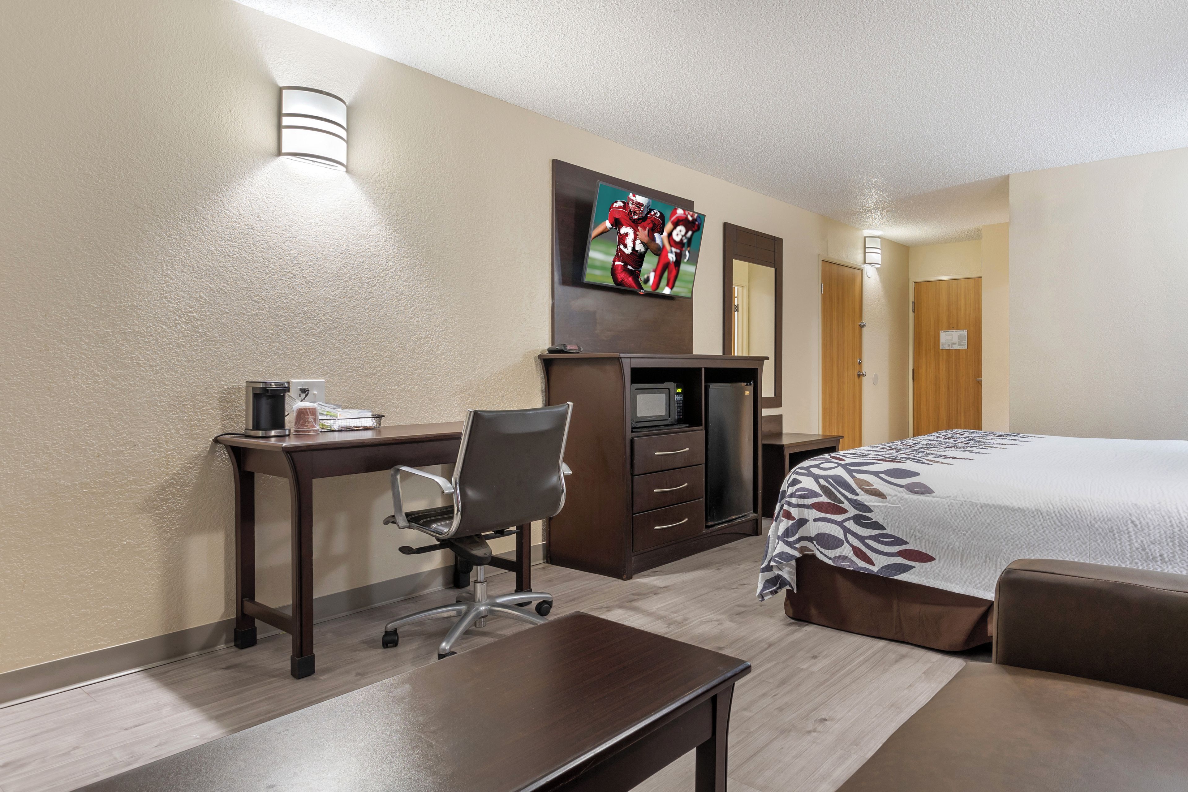 A hotel room with a bed, a desk with a chair, a television mounted on the wall, and a microwave.