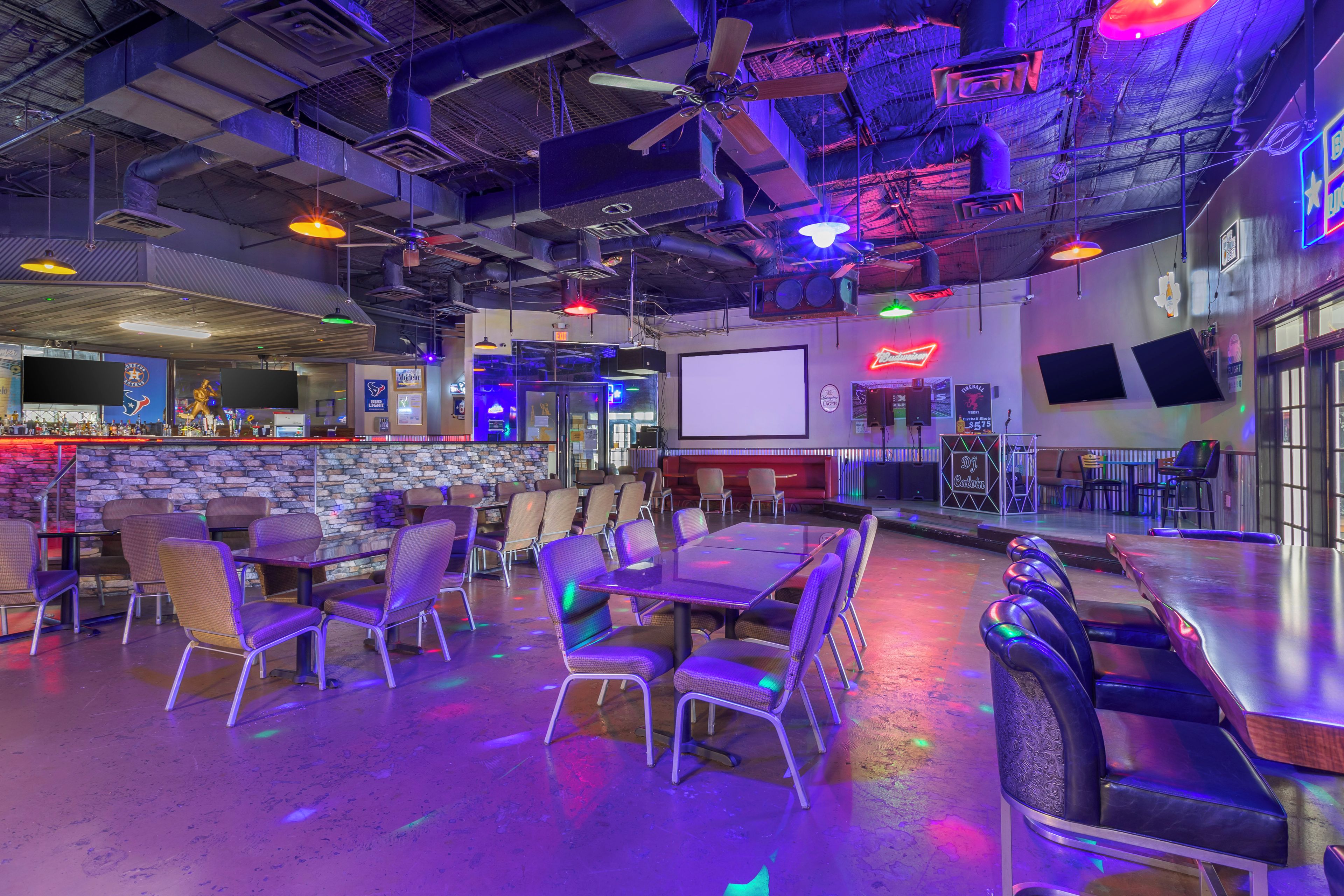 Un bar interior con mesas, sillas, un escenario y una pantalla.