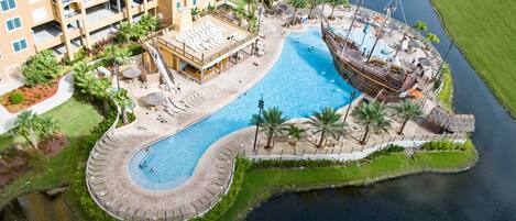 An aerial view of a resort with a large central pool, a ship-shaped water feature, and a surrounding landscaped area with palm trees and a small dock.