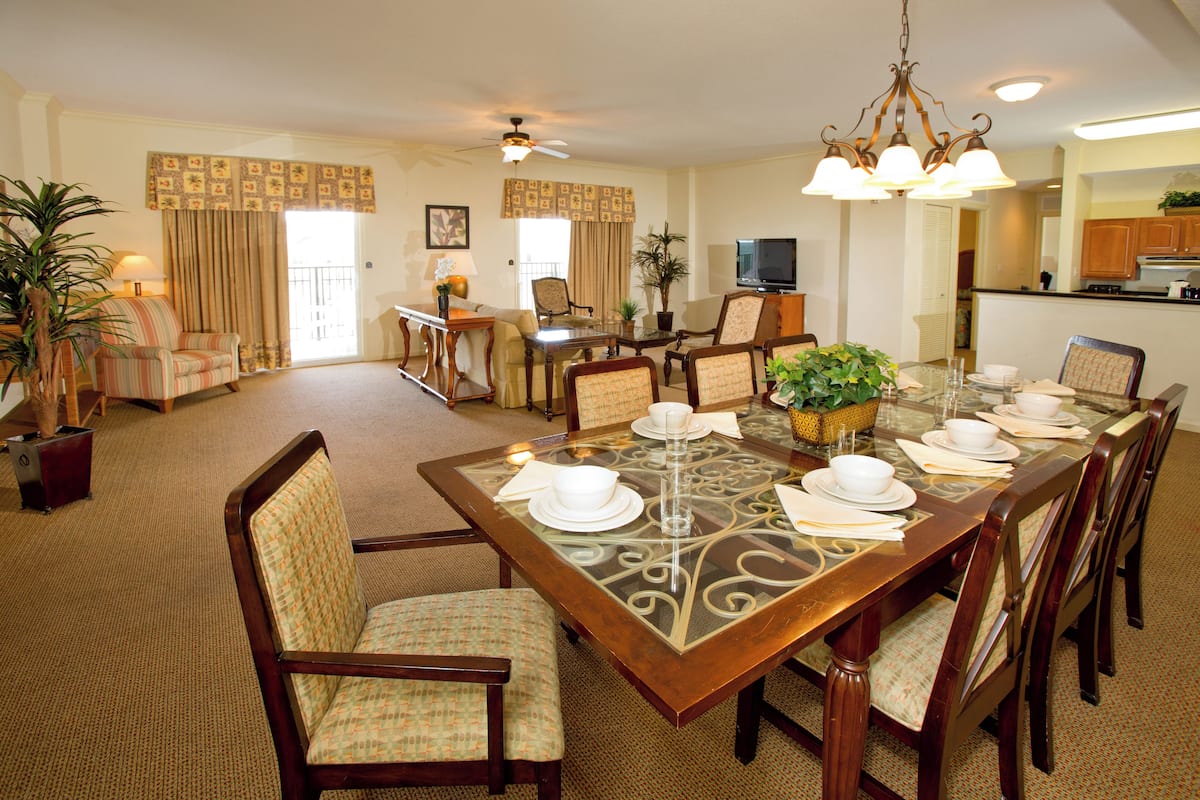 A dining area with a large table set for six, surrounded by chairs, in a well-lit room with a ceiling fan and a kitchen in the background.