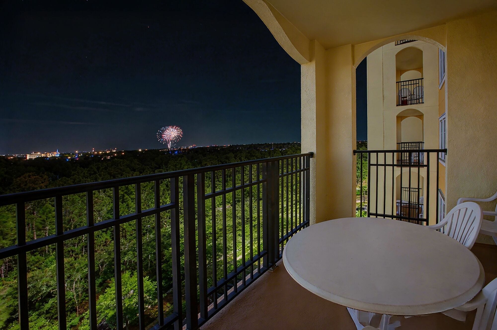 Een balkon met een tafel en stoelen, met uitzicht op een stadsbeeld en verre vuurwerk.